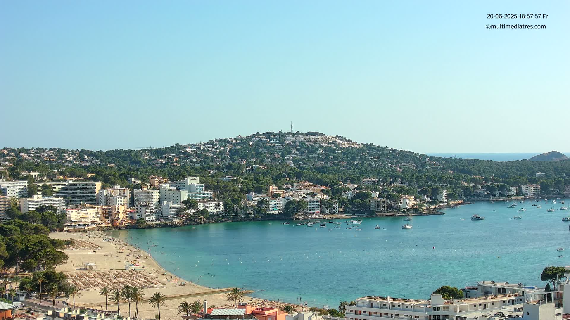 A coastal town with a sandy beach and calm, clear turquoise water sits below a hill densely covered with buildings and trees under a clear blue sky.