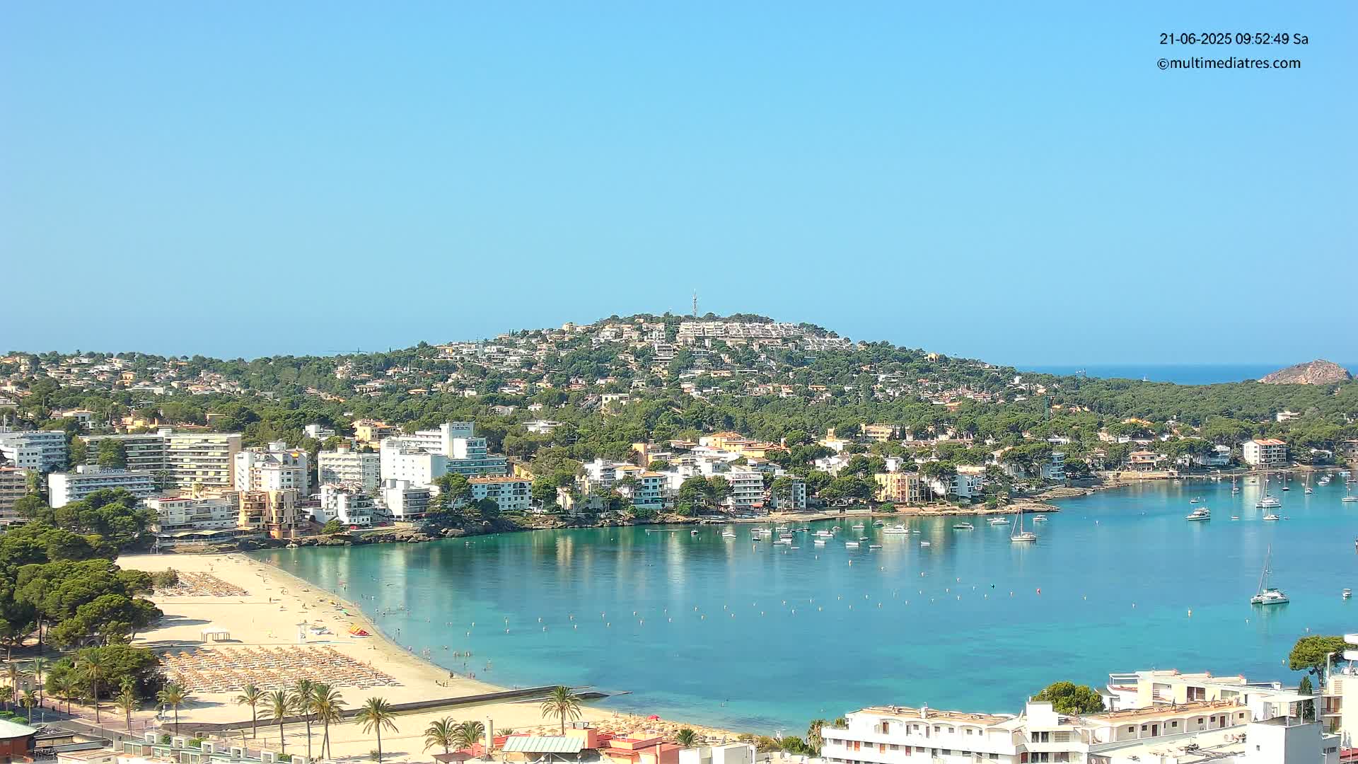 A sunny coastal town with a sandy beach and calm, turquoise water, nestled against a hill covered in buildings and lush greenery.