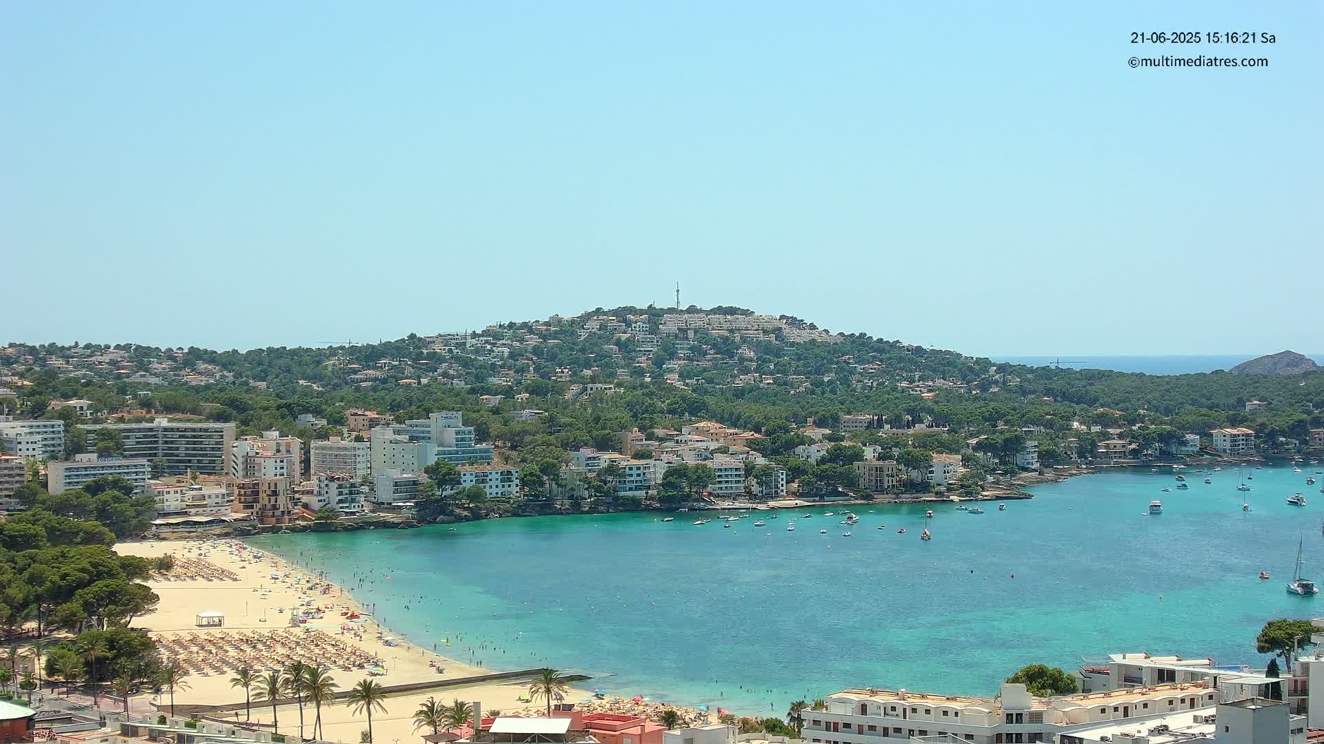 A sandy beach with sunbathers lines a turquoise bay filled with boats, nestled below a hill densely covered with buildings and trees under a clear, sunny sky.