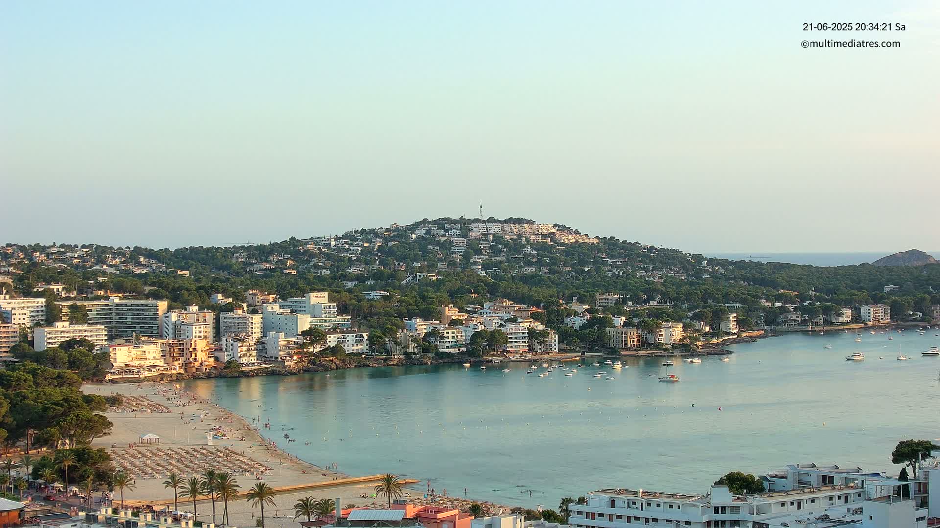 A coastal town nestled against a verdant hill overlooking a calm, turquoise bay with a sandy beach dotted with sunbathers under a clear, light-blue sky.
