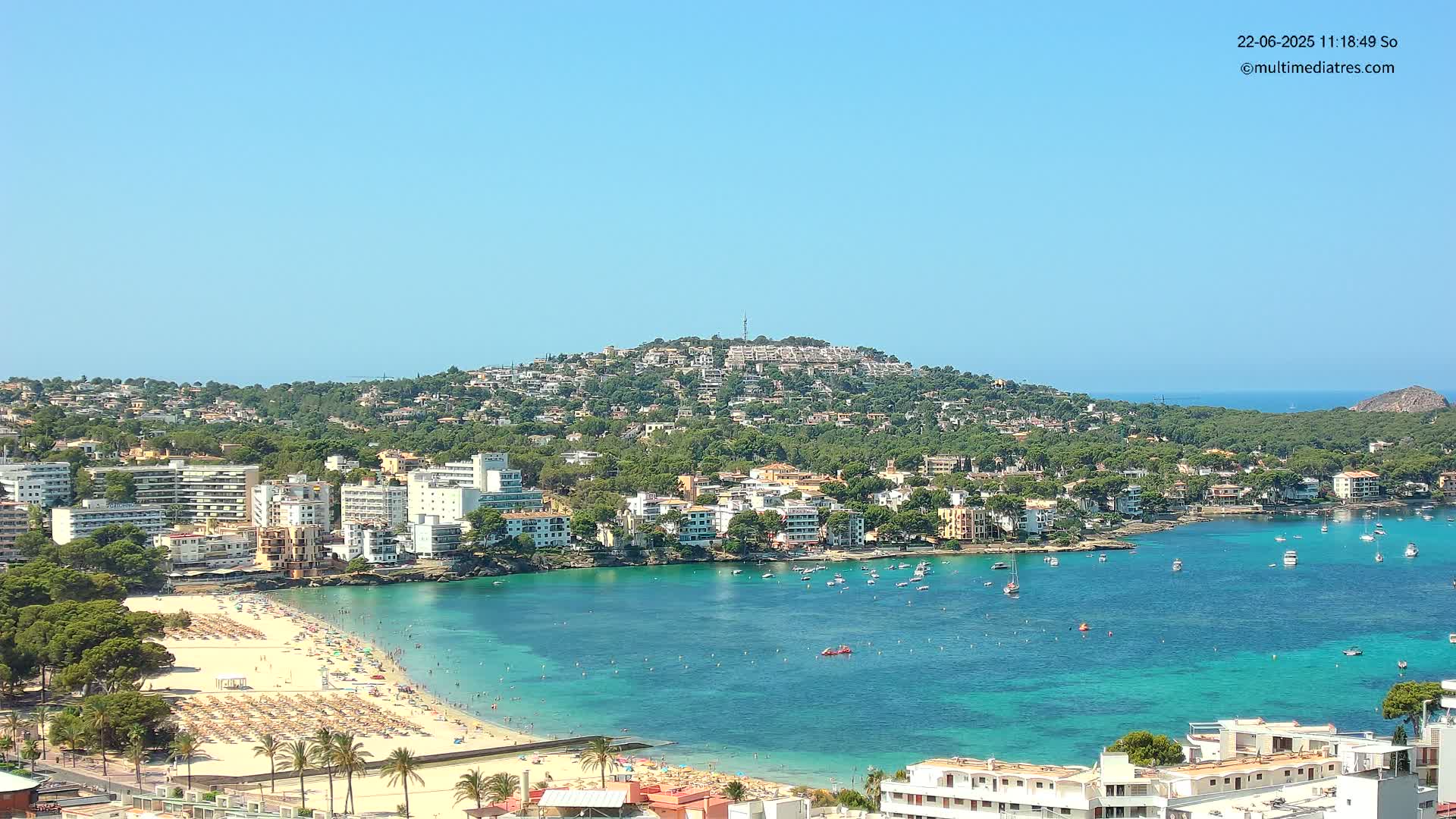 A coastal town with a sandy beach, turquoise water, and numerous buildings nestled on a hillside under a clear blue sky.