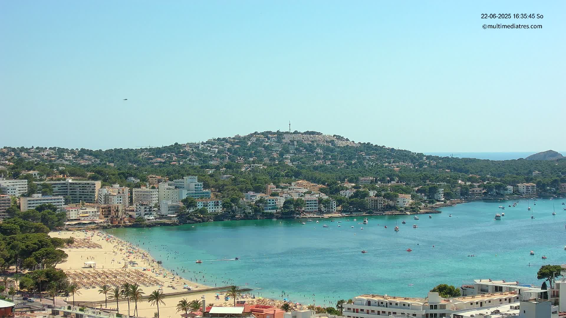 A clear, sunny day reveals a coastal town nestled on a bay, with a hill covered in buildings and trees in the background, and a sandy beach crowded with people in the foreground.