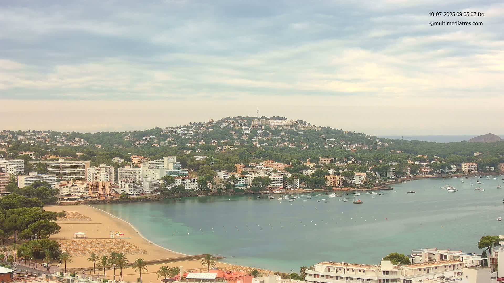 A coastal town with a sandy beach and calm bay is nestled beneath a hill covered in buildings and greenery under a cloudy sky.