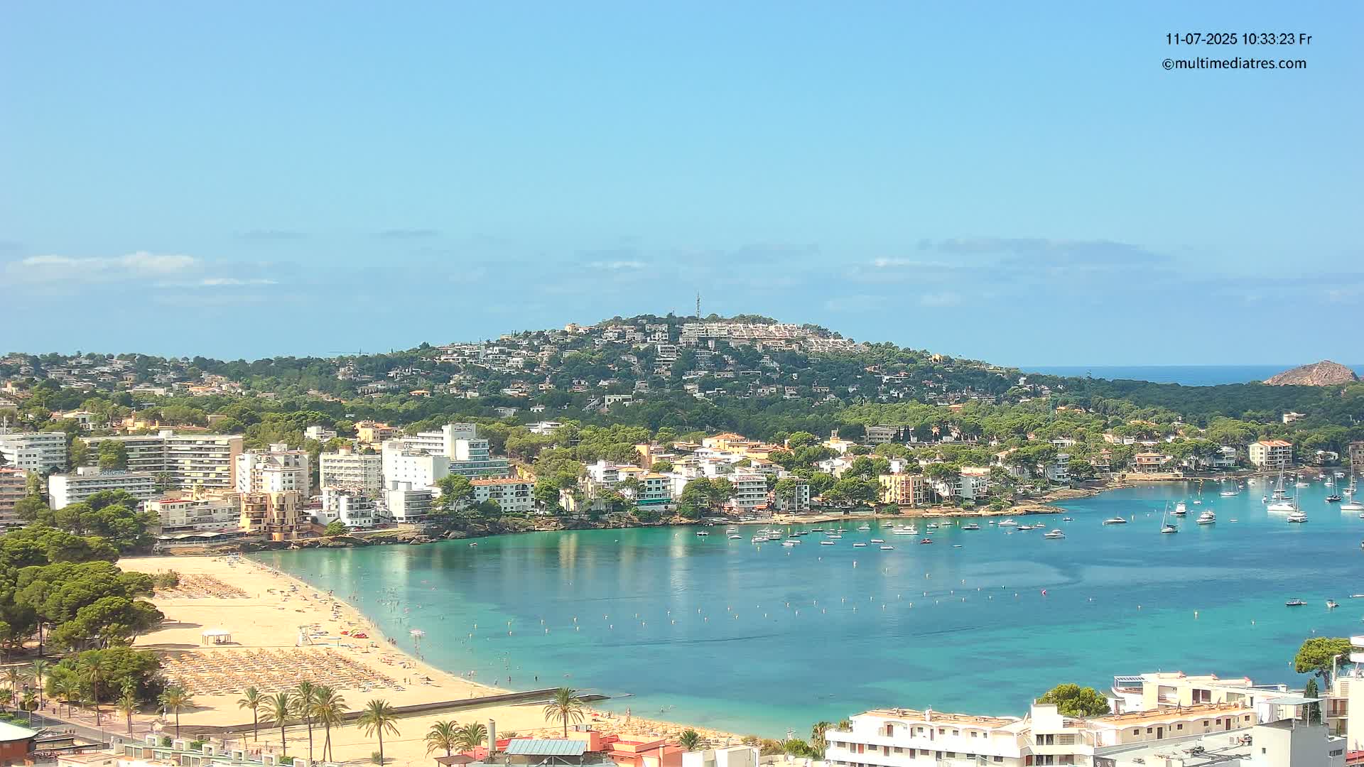 A sunny coastal town with a sandy beach, calm turquoise water filled with boats, and a hill covered in buildings and lush greenery in the background.