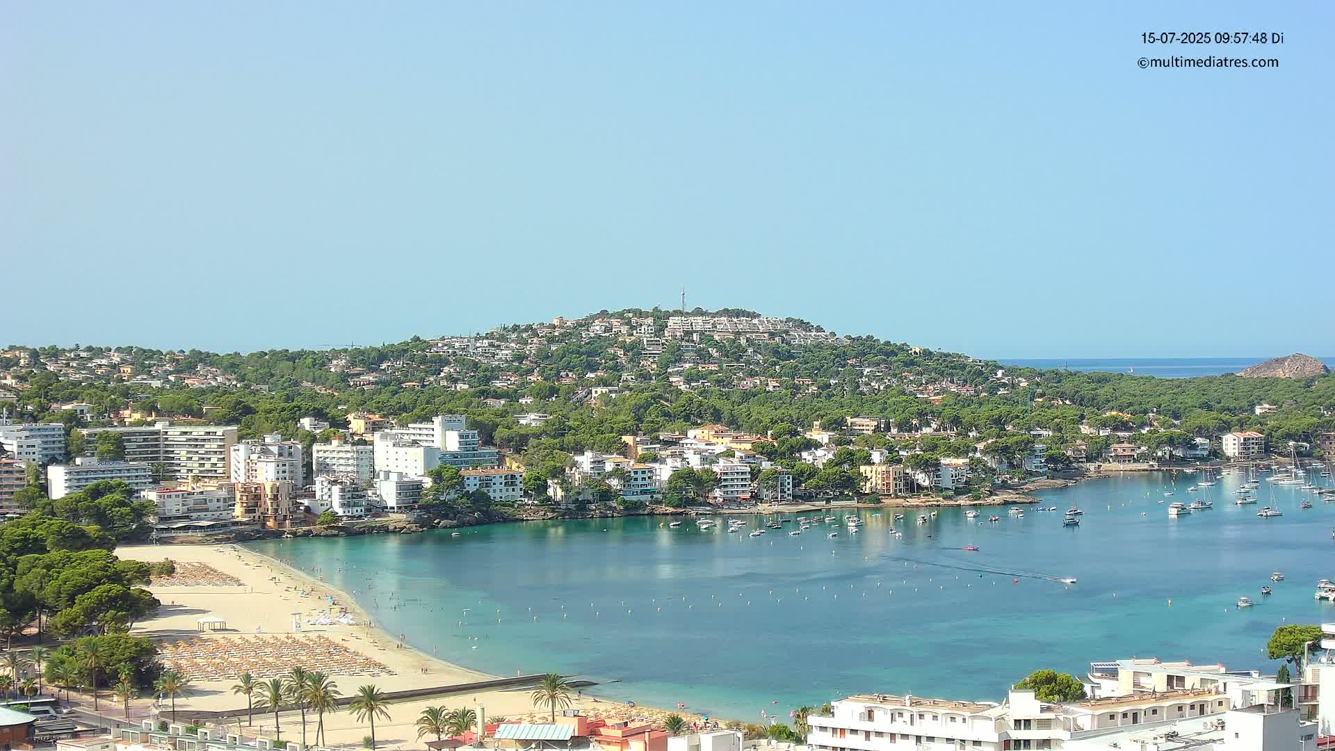 A coastal town nestled into a hillside, featuring a sandy beach, calm blue water filled with boats, and clear sunny skies.