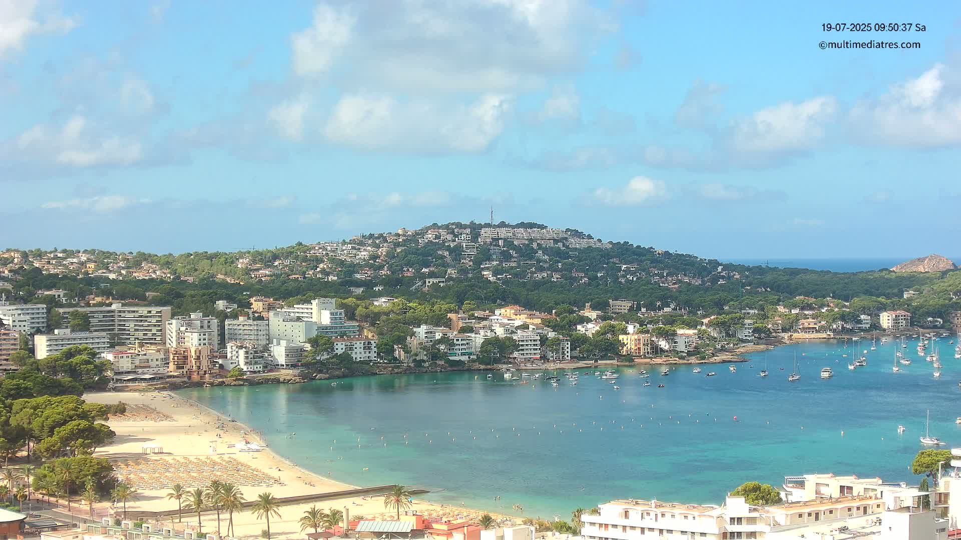 A sunny coastal town with a sandy beach, calm turquoise water filled with boats, and buildings nestled on a hillside under a partly cloudy sky.