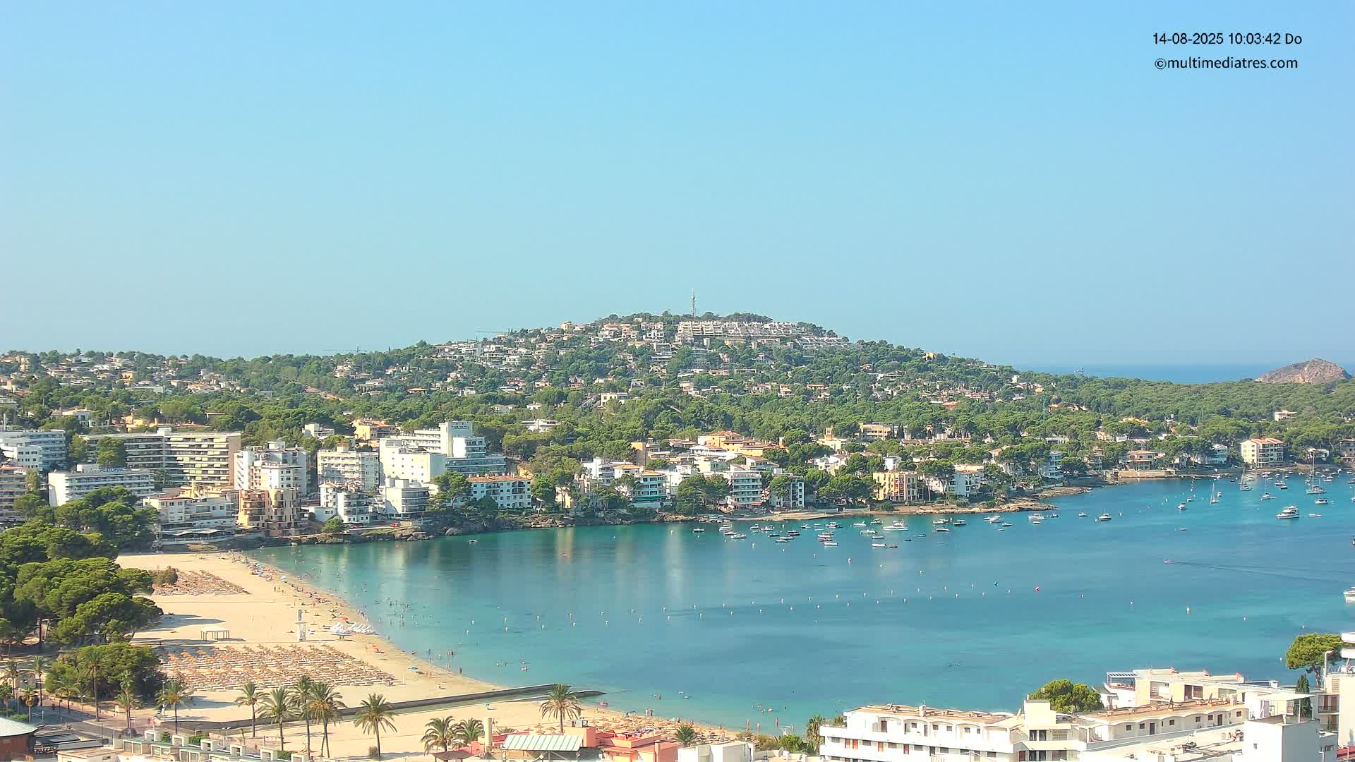 A coastal town with a sandy beach and calm, clear turquoise water is nestled beneath a hill covered in buildings and lush green vegetation, under a clear blue sky.