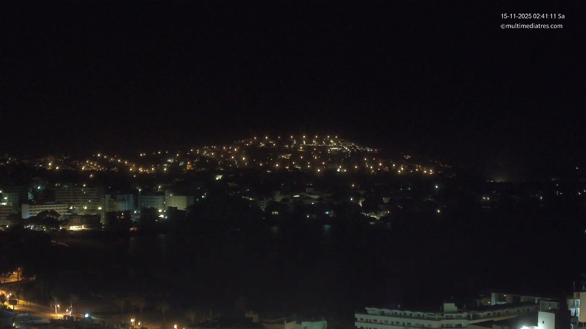 A panoramic night view captures a town sprawling across a dark hillside and along a calm body of water, brightly illuminated by numerous scattered lights under a clear, starless sky.