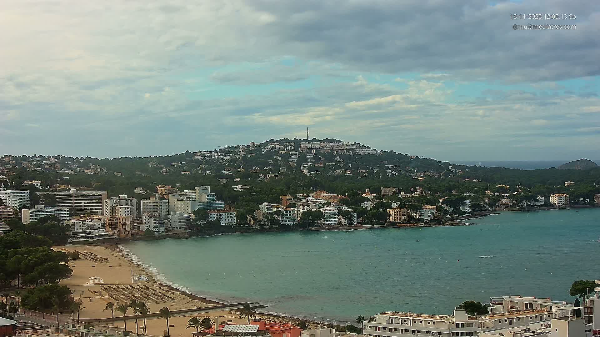 A panoramic view reveals a coastal town with a sandy beach and a turquoise bay, flanked by numerous buildings extending up a tree-covered hill topped with a tower, all under a cloudy sky.