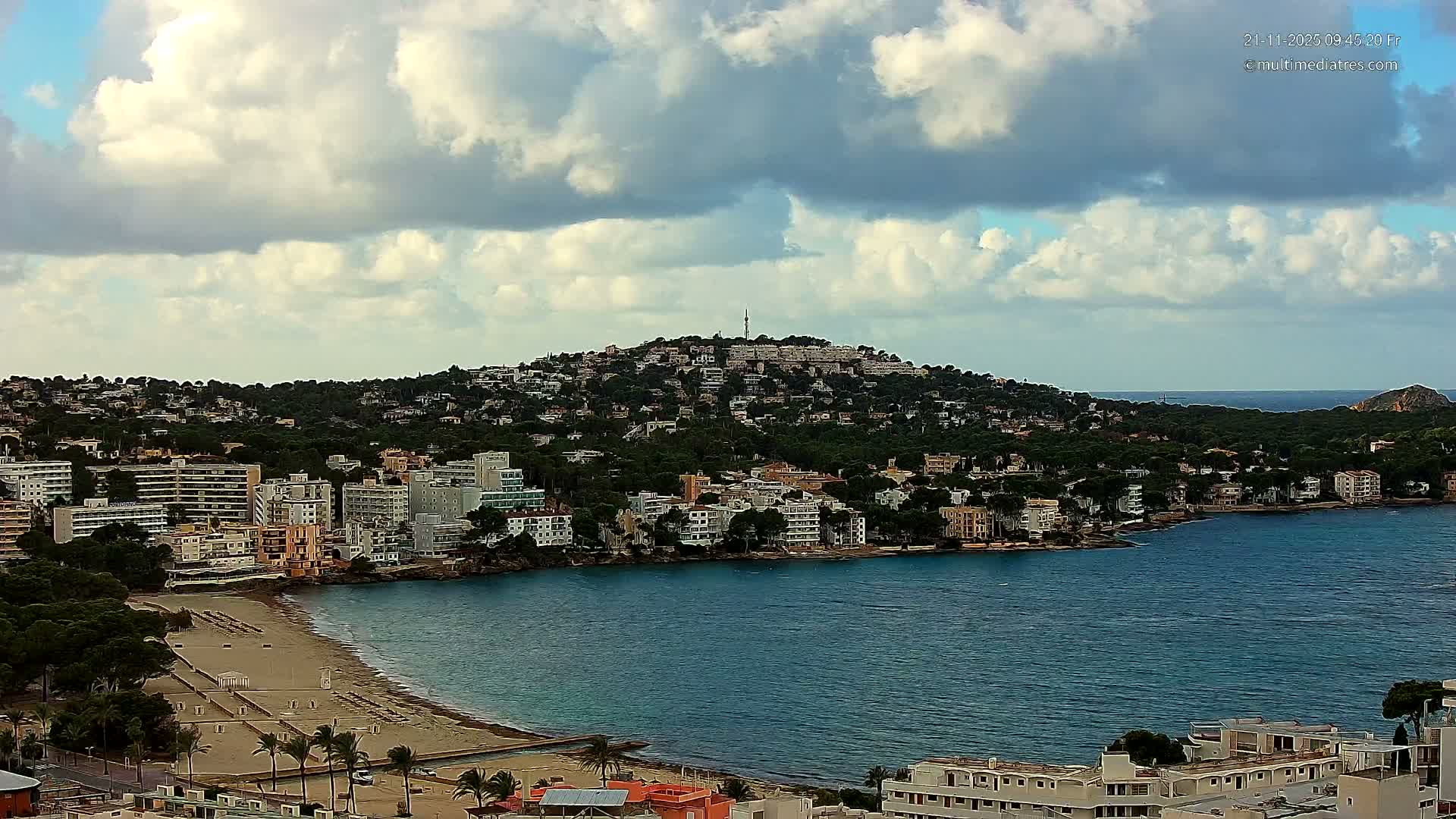 An expansive view captures a coastal town with a sandy beach and numerous buildings along a bay, extending up a densely wooded hillside topped by a communication tower, all under a partly cloudy sky.
