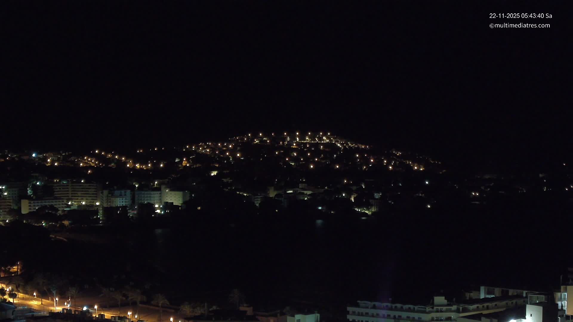 A nighttime panoramic view reveals a coastal town with numerous illuminated buildings, streetlights along a waterfront road, and a distant hillside dotted with more lights under a clear, dark sky.