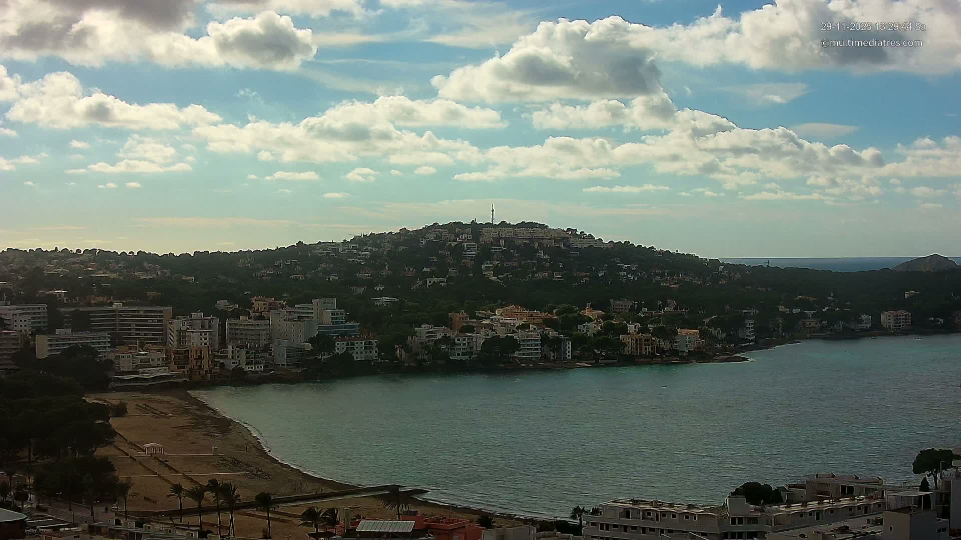An expansive view reveals a coastal town featuring a sandy beach curving into a bay, bordered by numerous buildings and a prominent, tree-covered hill crowned with a communication tower, all under a partly cloudy sky.