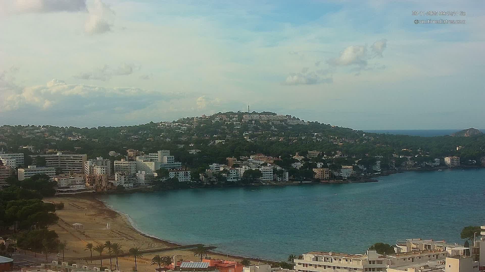 An aerial view reveals a scenic coastal town with a sandy beach and buildings bordering a calm bay, backed by a densely built-up, tree-covered hill, under a sky with scattered clouds.
