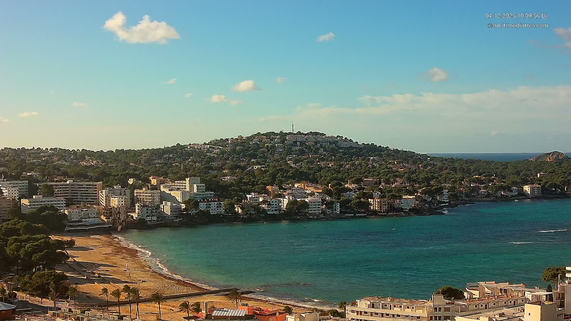 The image captures a sunny coastal town with numerous buildings nestled among trees on a hill overlooking a sandy beach and a bay with turquoise waters, under a clear blue sky with a few scattered clouds.