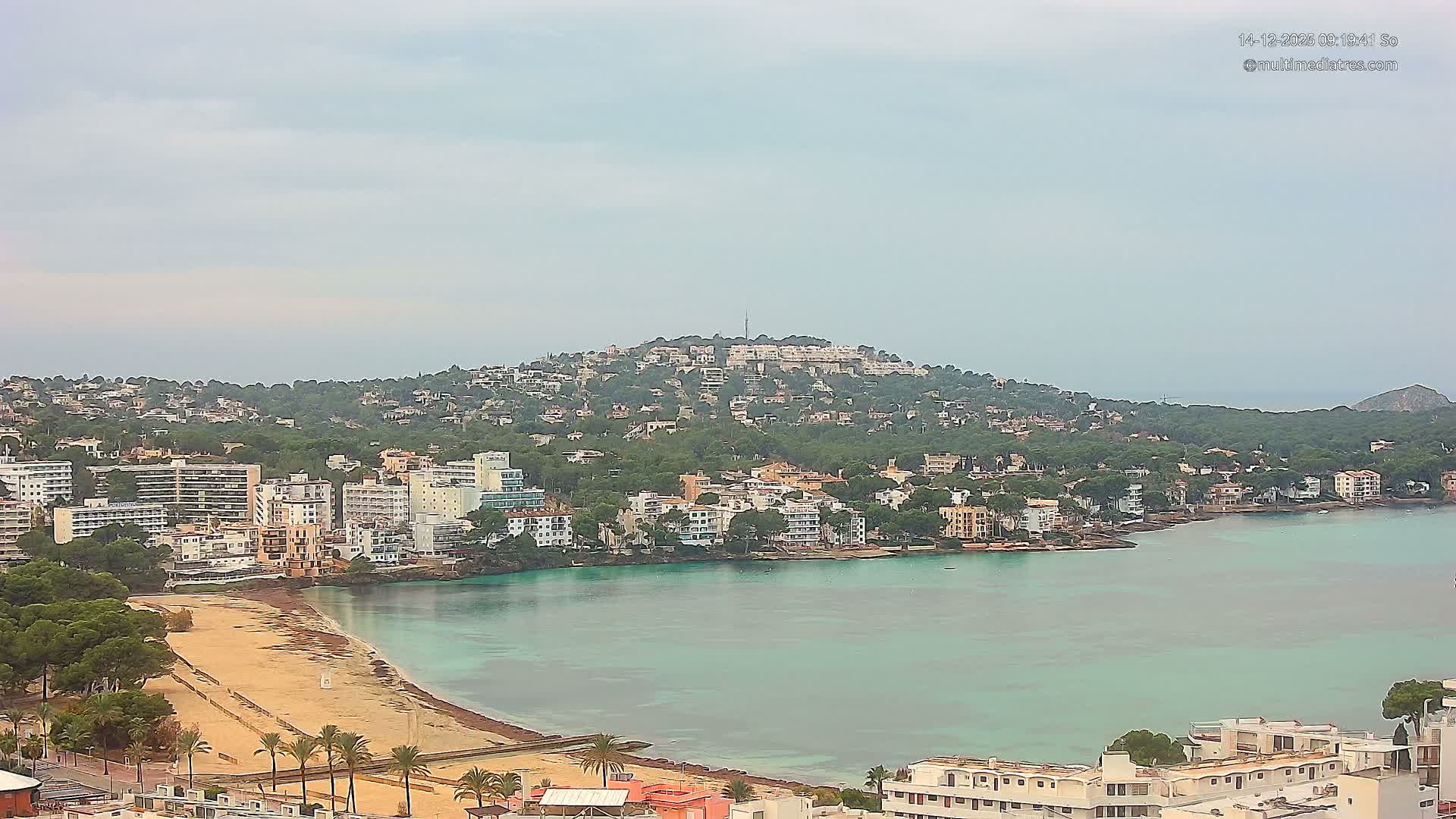 The image captures a sunny coastal town with numerous buildings nestled among trees on a hill overlooking a sandy beach and a bay with turquoise waters, under a clear blue sky with a few scattered clouds.