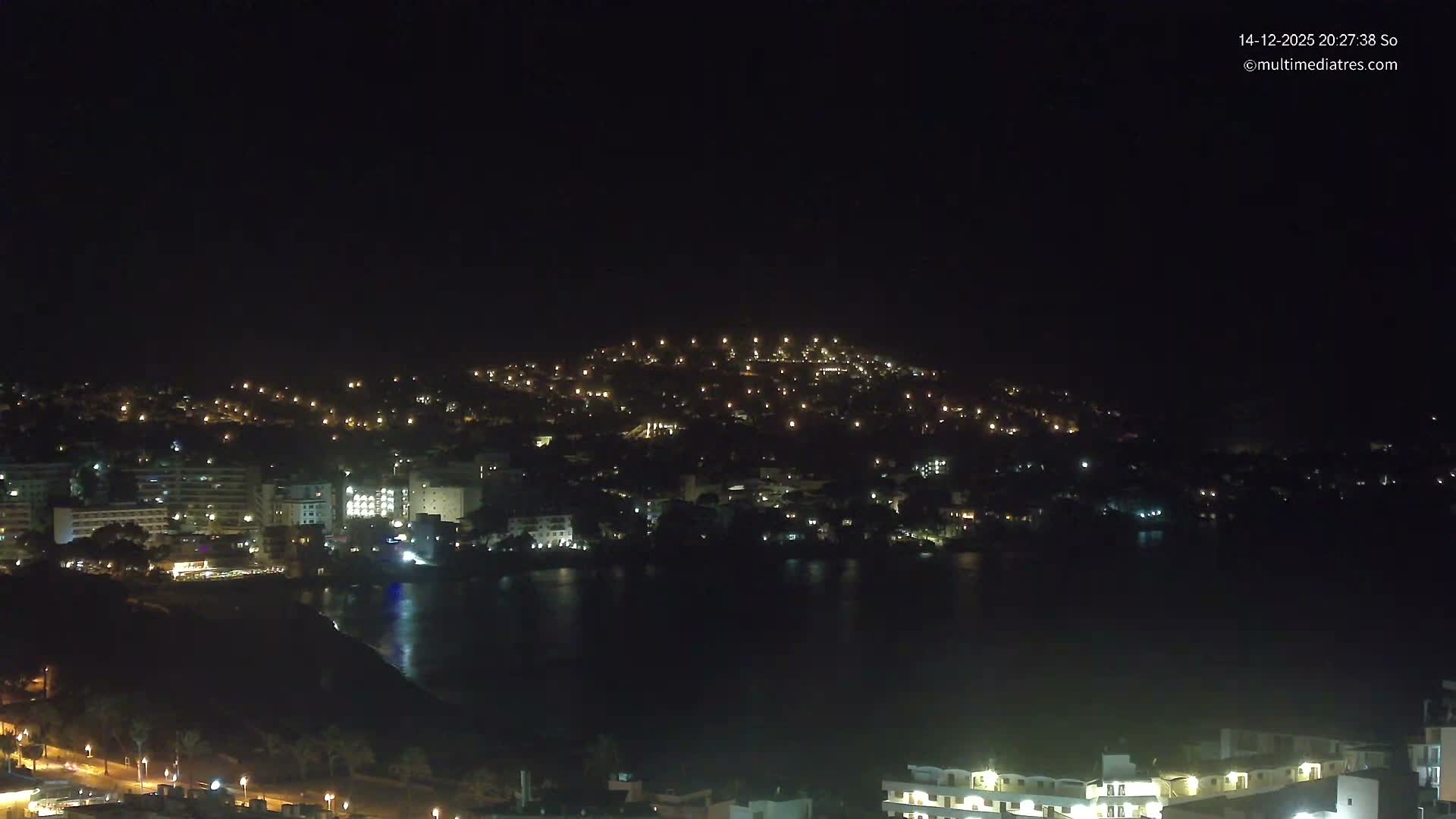 The image captures a sunny coastal town with numerous buildings nestled among trees on a hill overlooking a sandy beach and a bay with turquoise waters, under a clear blue sky with a few scattered clouds.