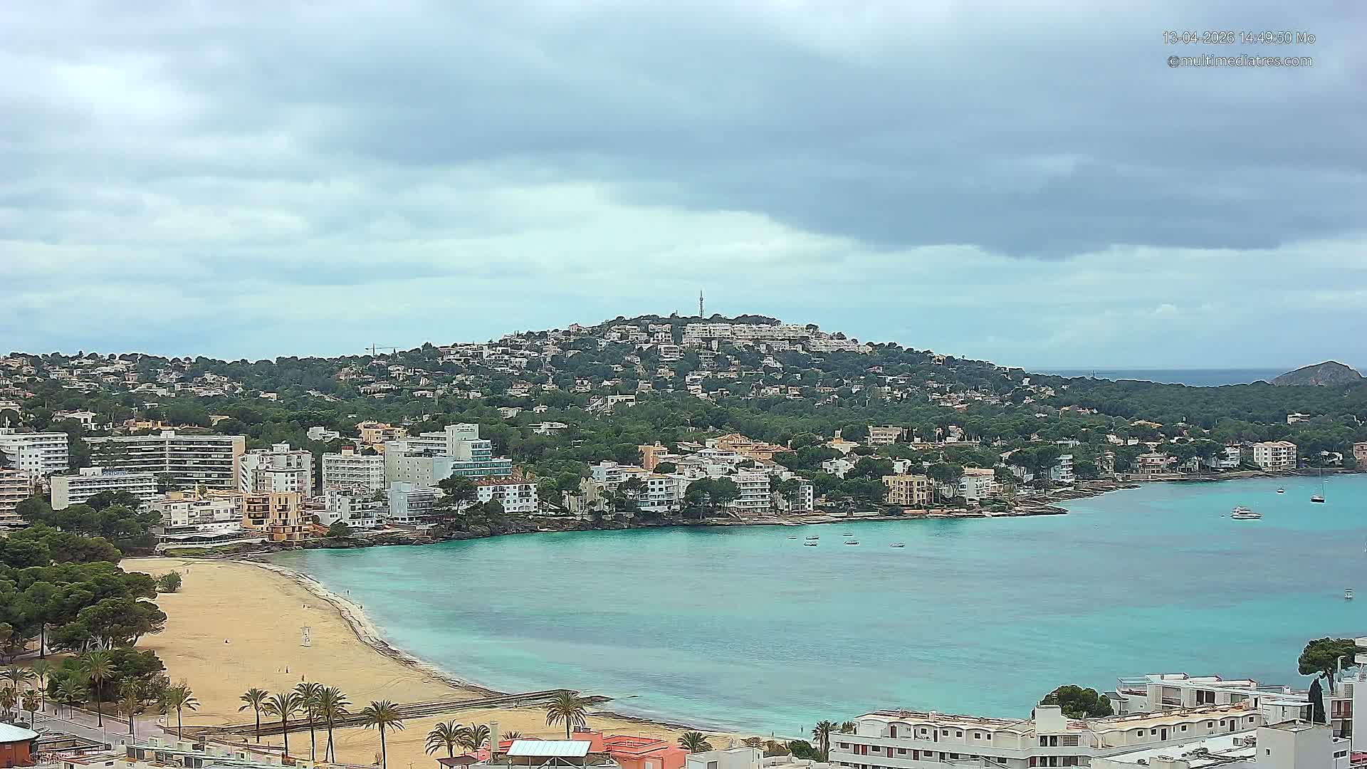 The image captures a sunny coastal town with numerous buildings nestled among trees on a hill overlooking a sandy beach and a bay with turquoise waters, under a clear blue sky with a few scattered clouds.