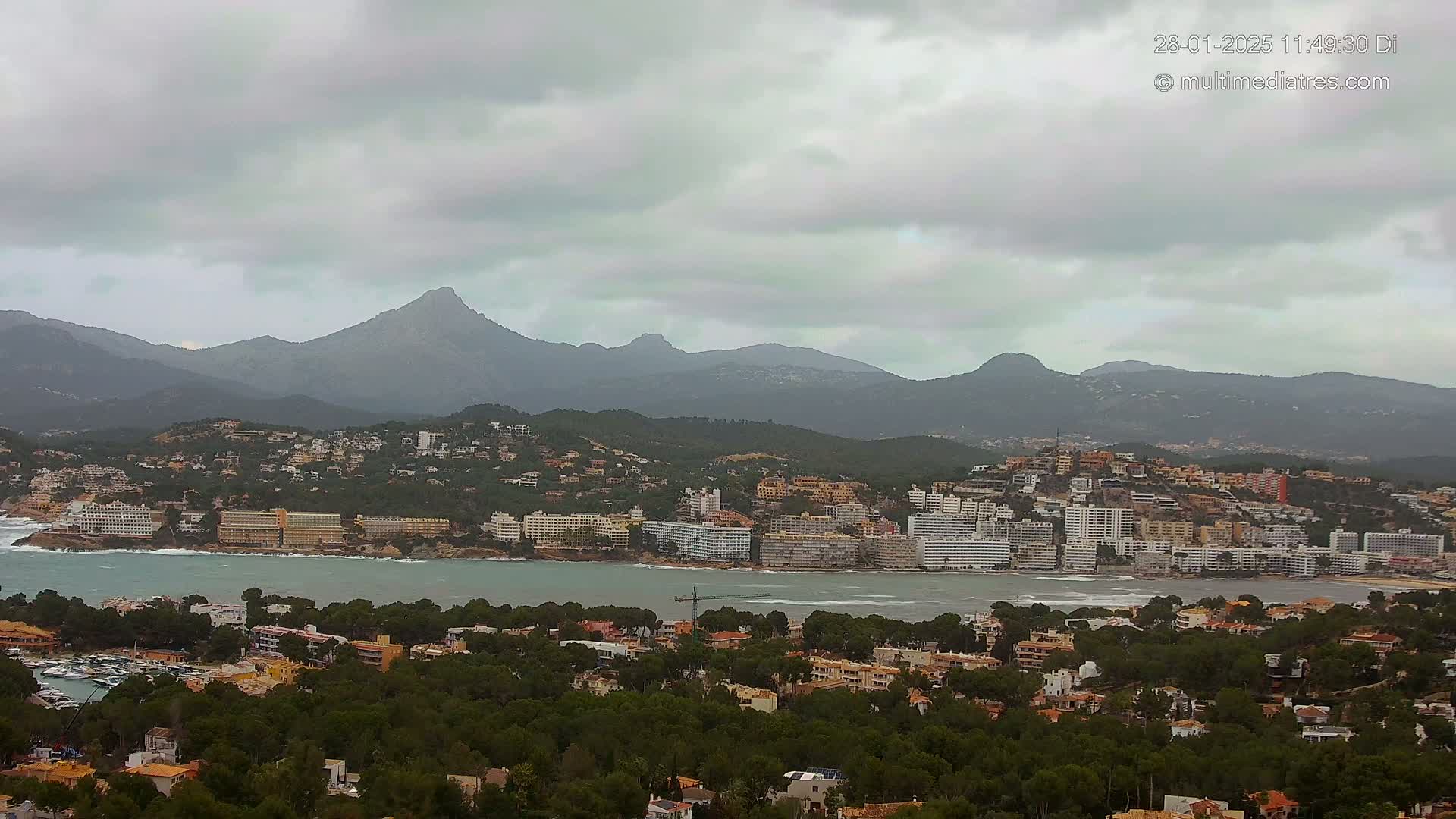 A coastal town nestled among green hills and mountains under a cloudy sky, with a body of water separating the town from a line of hotels.