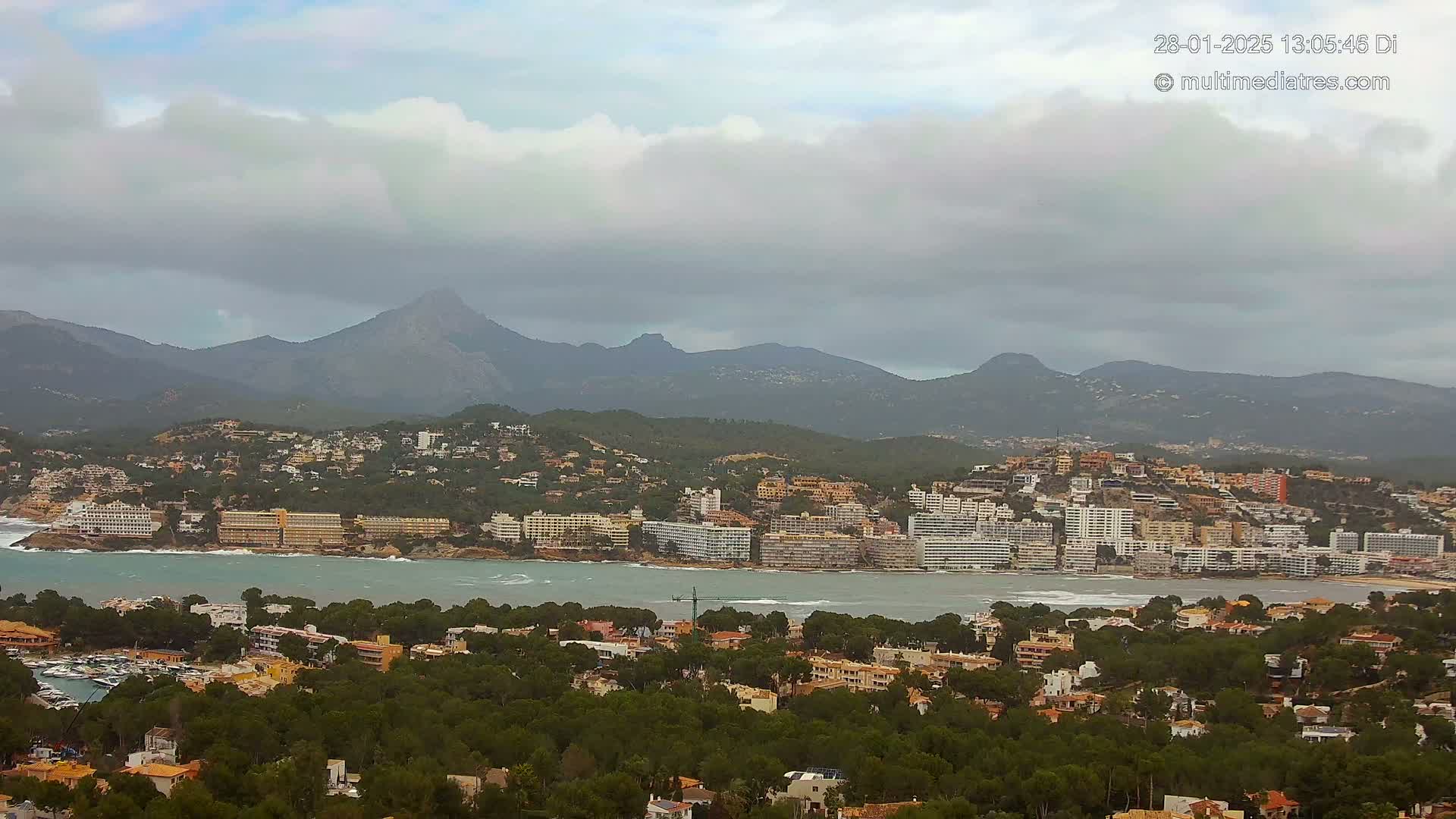 A coastal town with many buildings nestles beside a calm bay, overlooked by a mountainous range under a cloudy sky.