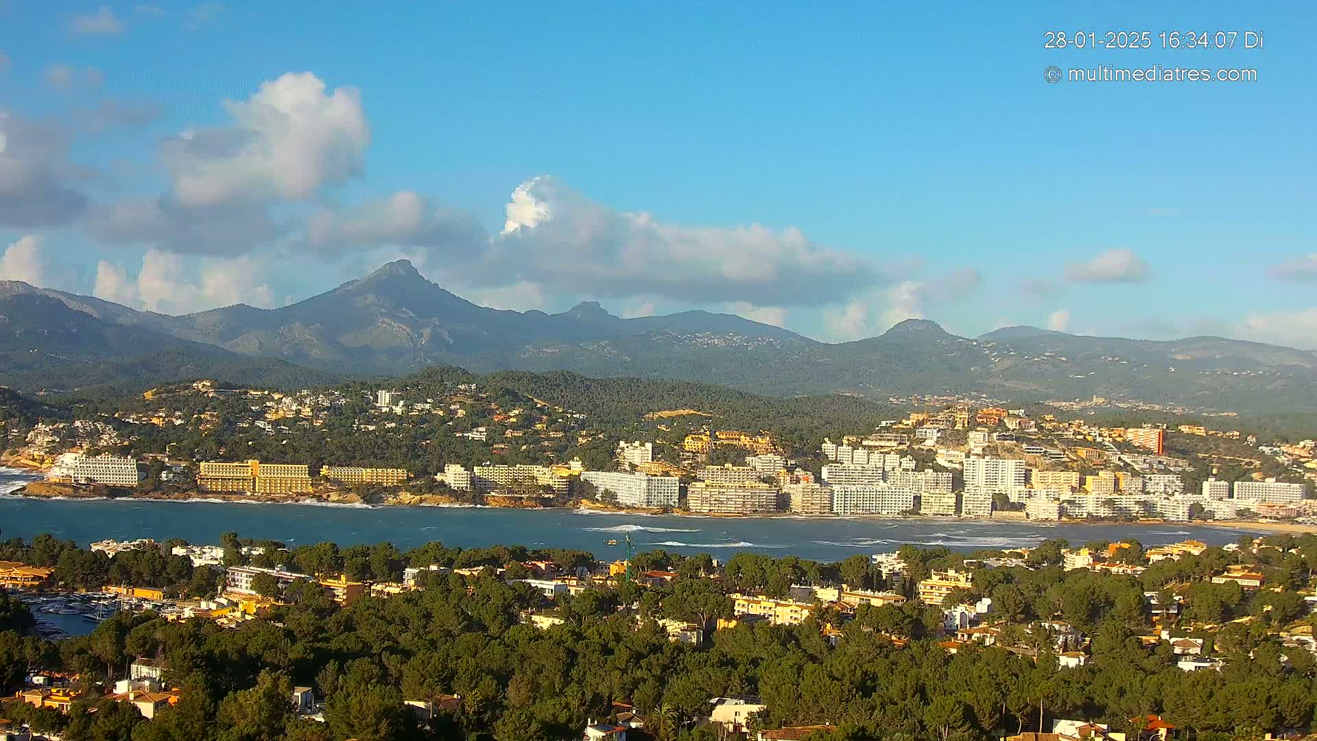 A coastal town nestled between a calm bay and a mountain range under a partly cloudy, sunny sky.