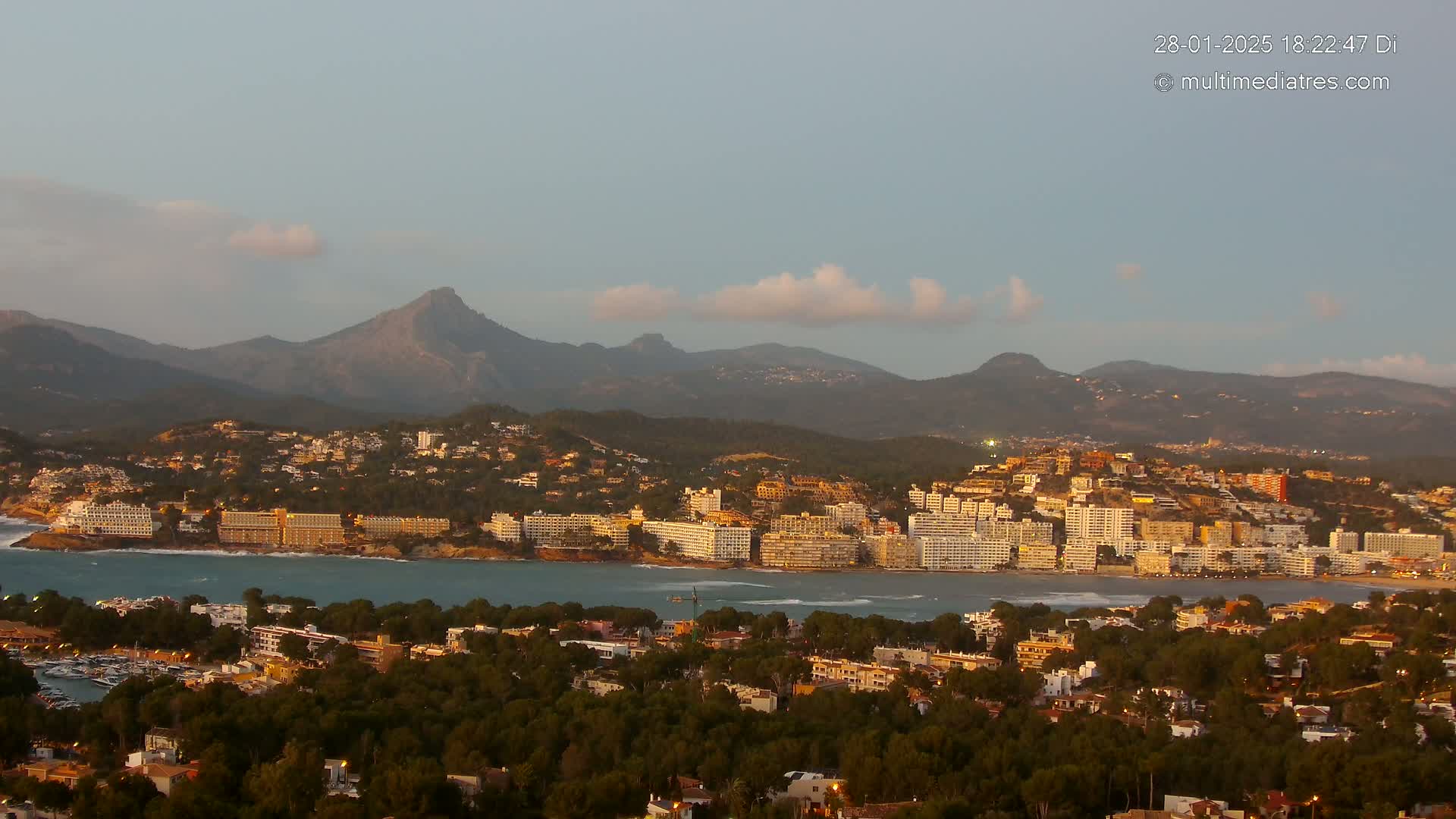 A coastal town nestled between a calm bay and a mountain range under a partly cloudy twilight sky.