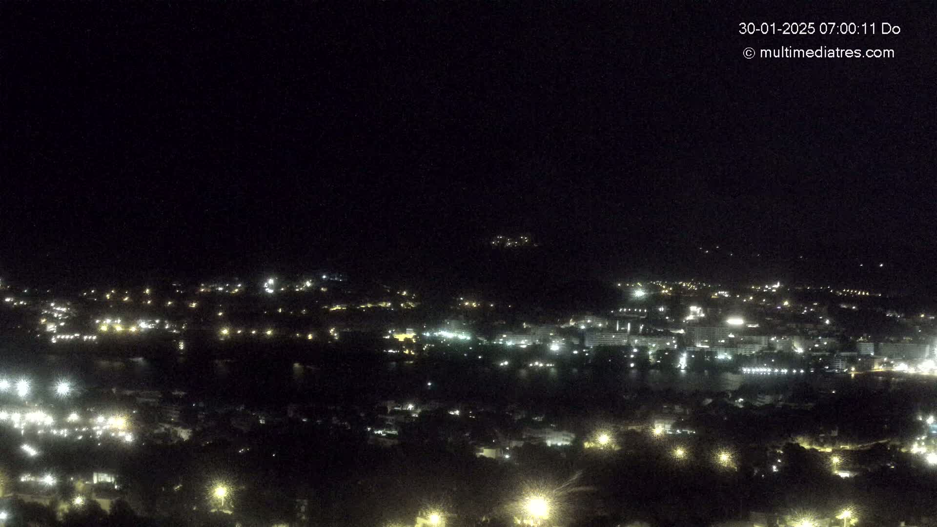 A nighttime, high-angle view shows a coastal town with scattered lights and a dark body of water.