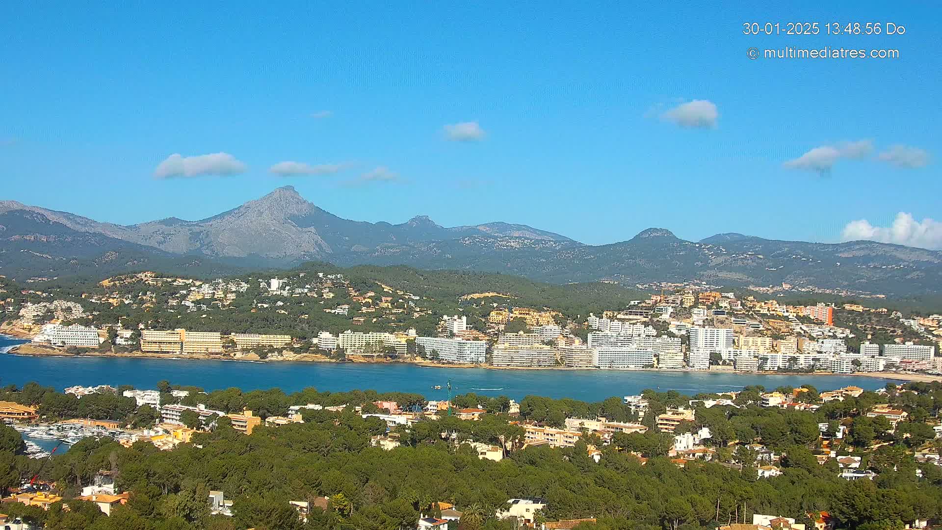 A coastal town with hotels and residential buildings is nestled beside a calm bay, backed by lush green hills and a mountain range under a clear blue sky with a few small clouds.