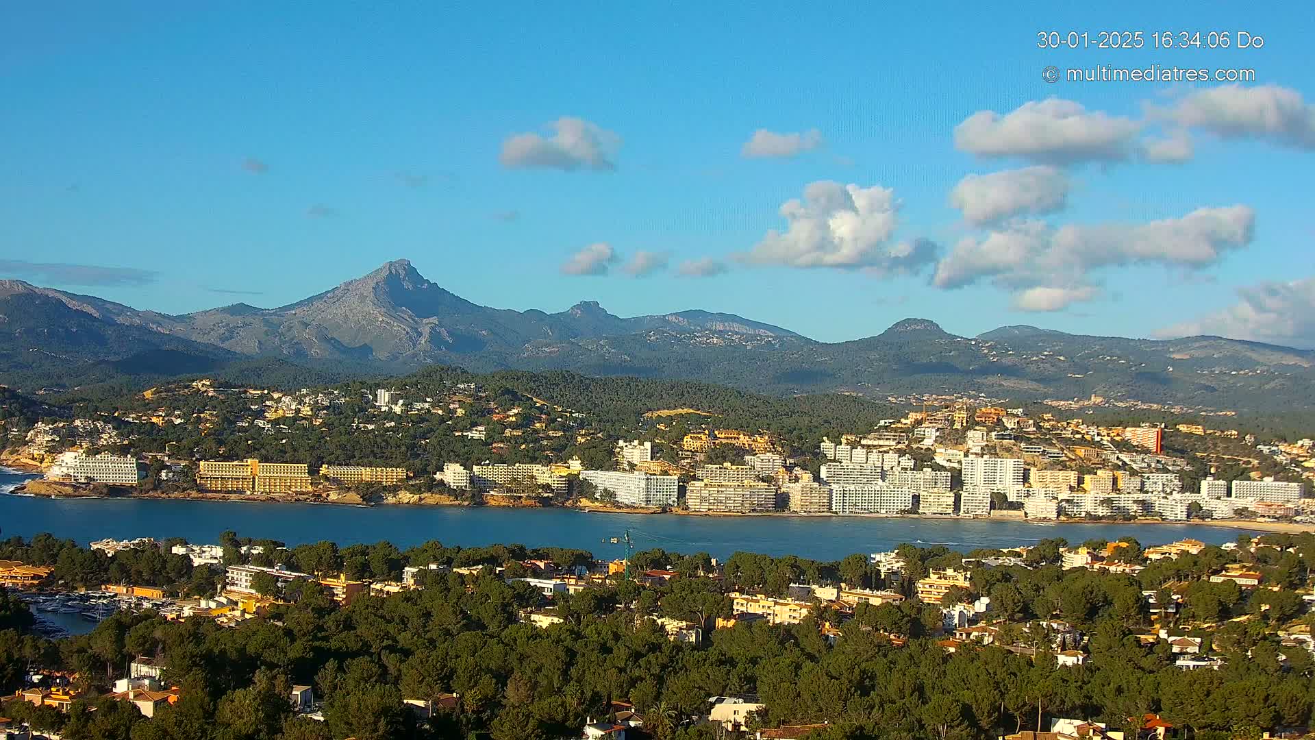 A coastal town with numerous hotels and buildings sits nestled beside a calm bay, with mountains rising in the background under a mostly clear, sunny sky.