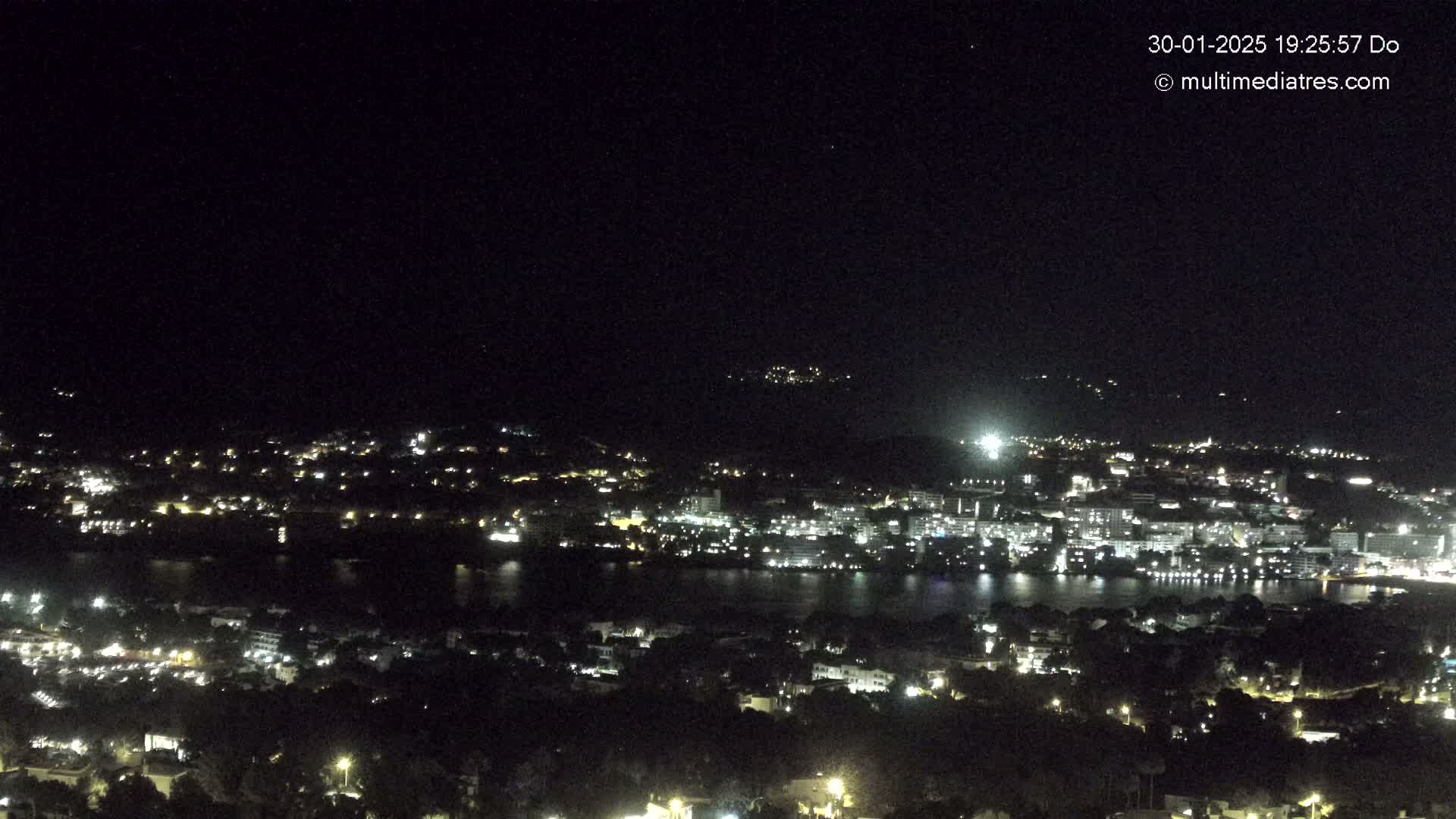 A nighttime, high-angle view shows a coastal town illuminated with many lights, situated next to a dark body of water, all under a clear, dark sky.