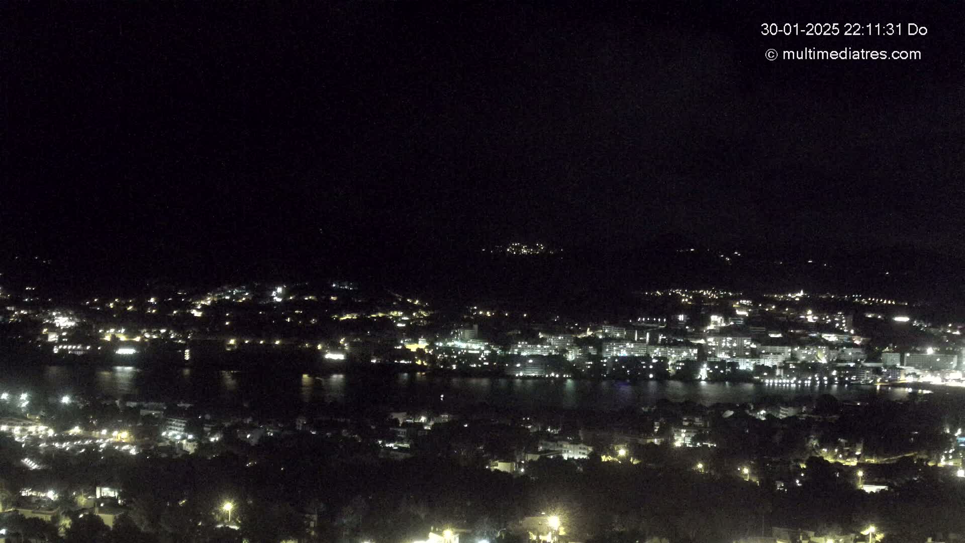 A nighttime, high-angle view shows a city and body of water illuminated by lights, with a dark sky above.