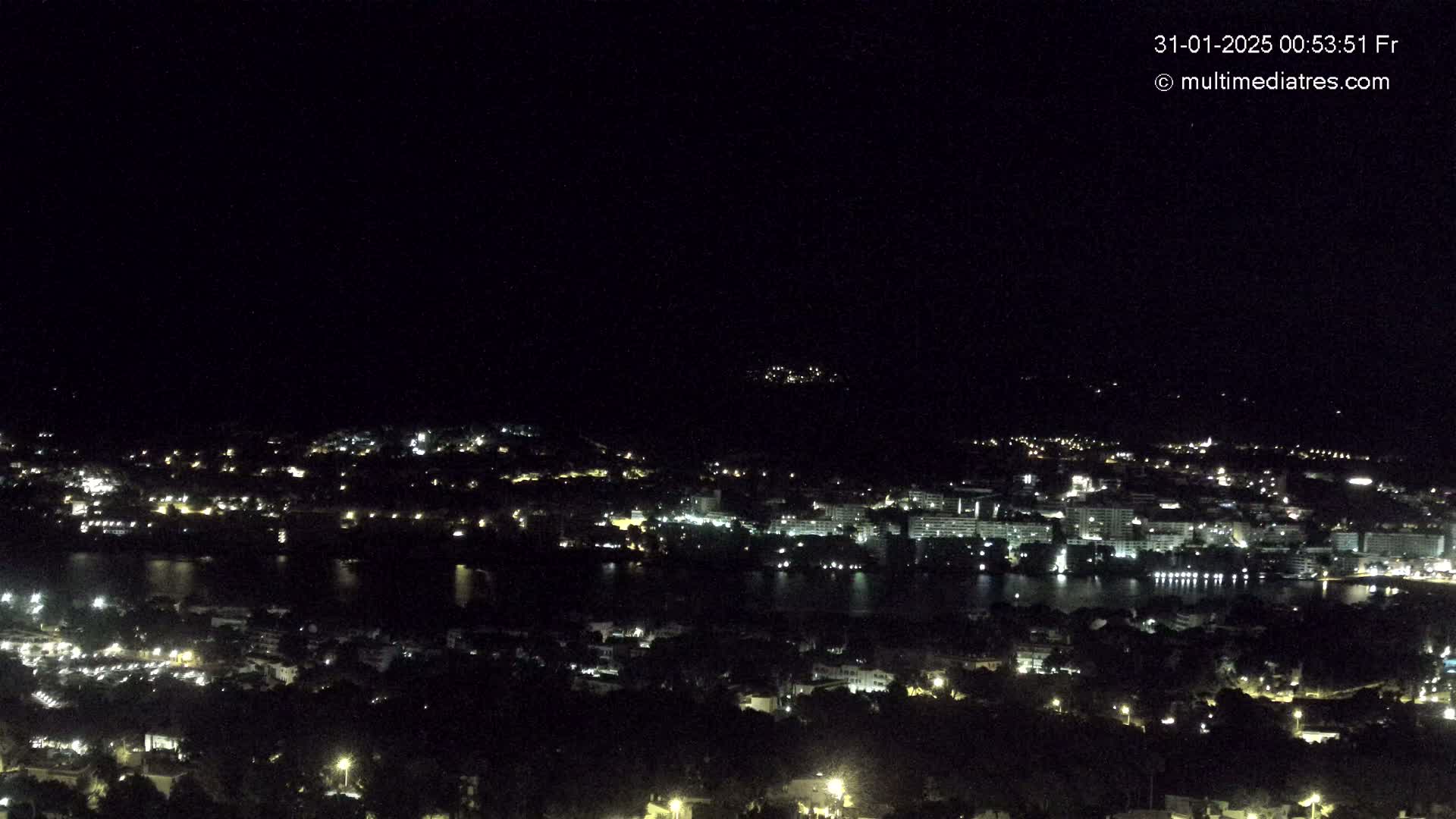A nighttime, aerial view of a coastal town with scattered lights illuminating buildings and a body of dark water.