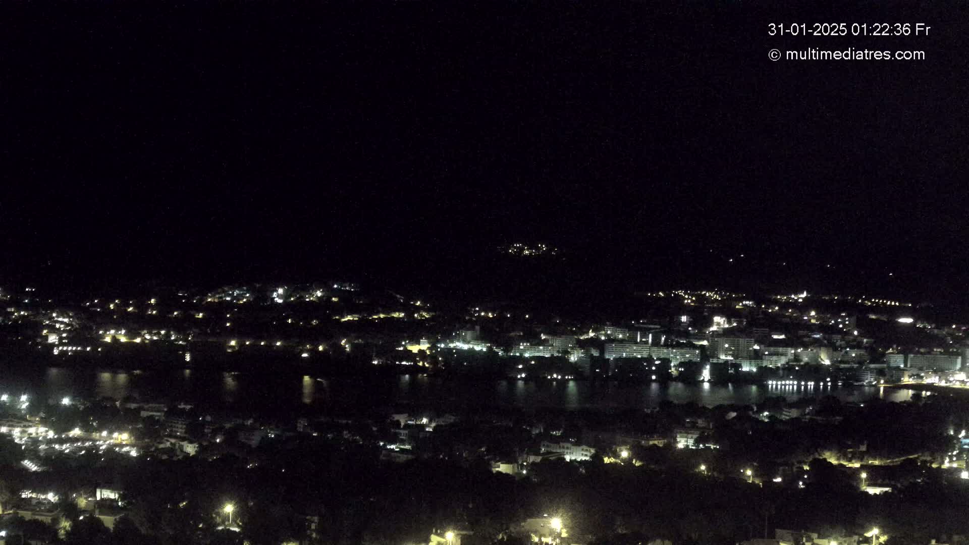 A nighttime, aerial view shows a city spread across a hillside and along a body of water, with scattered lights illuminating the landscape under a clear, dark sky.