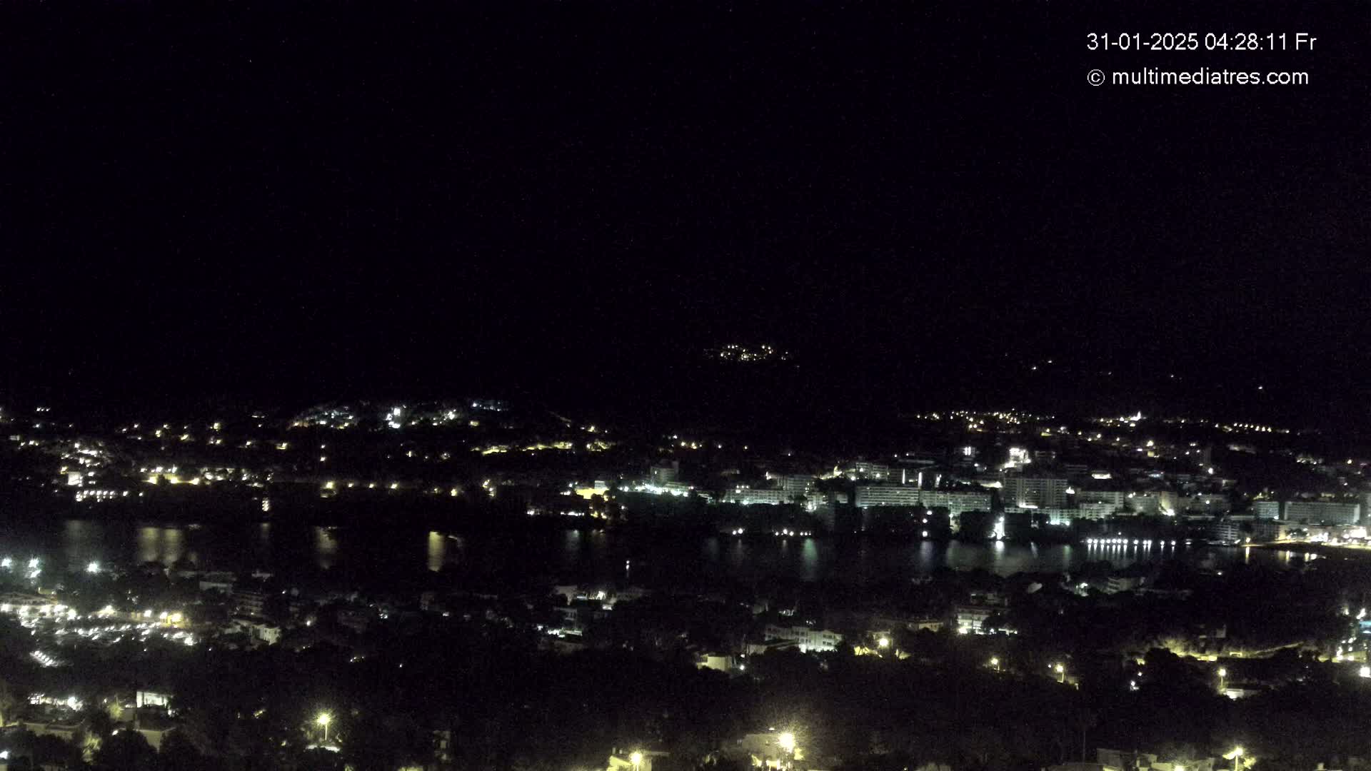 A nighttime, high-angle view shows a city with many lights beside a dark body of water, under a clear, dark sky.