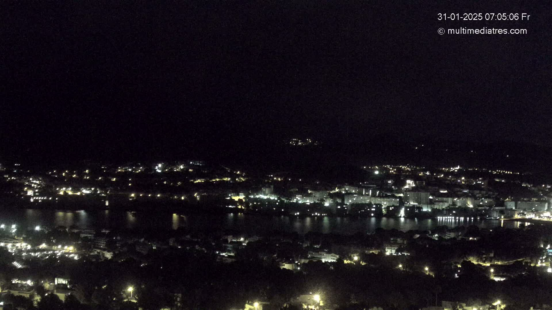 A nighttime, high-angle view shows a coastal town with its lights illuminating the dark landscape and a body of water.