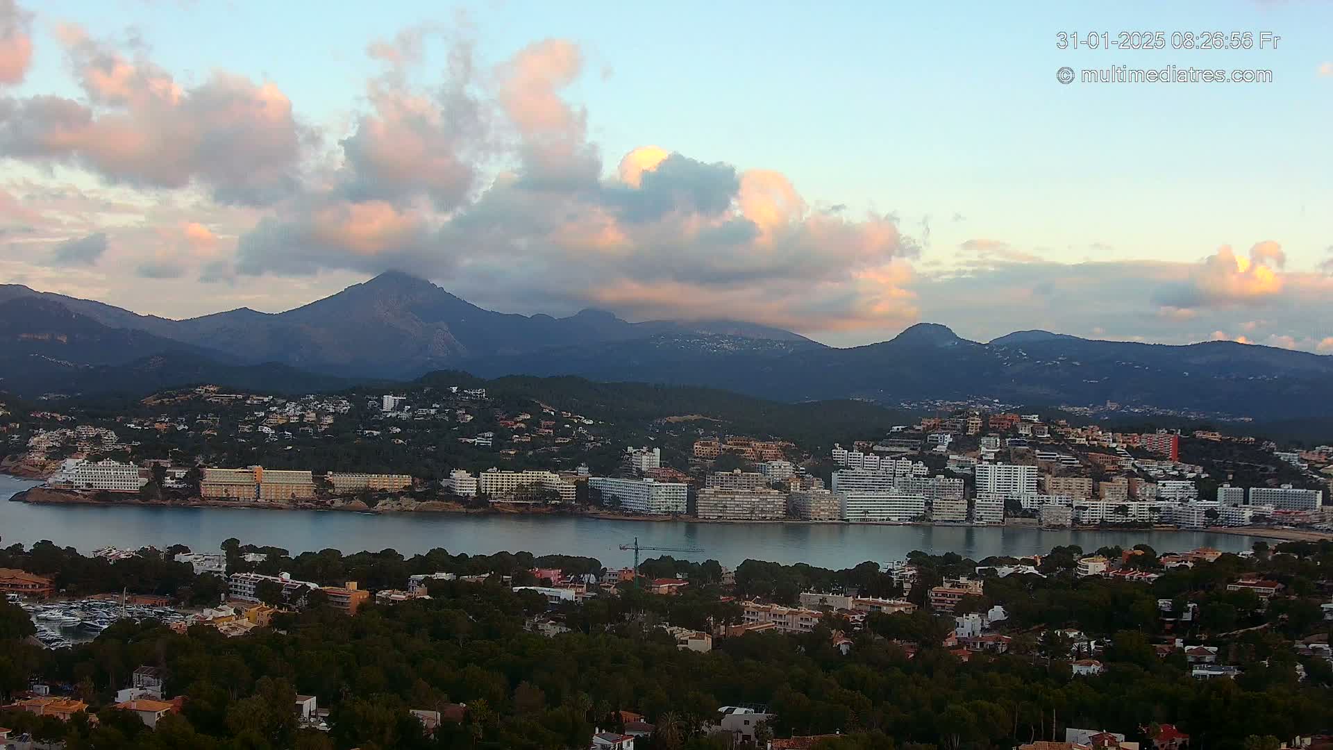 A coastal town nestled beside a calm bay, with hotels and buildings lining the waterfront, is overlooked by mountains under a partly cloudy, pinkish sky at dawn or dusk.