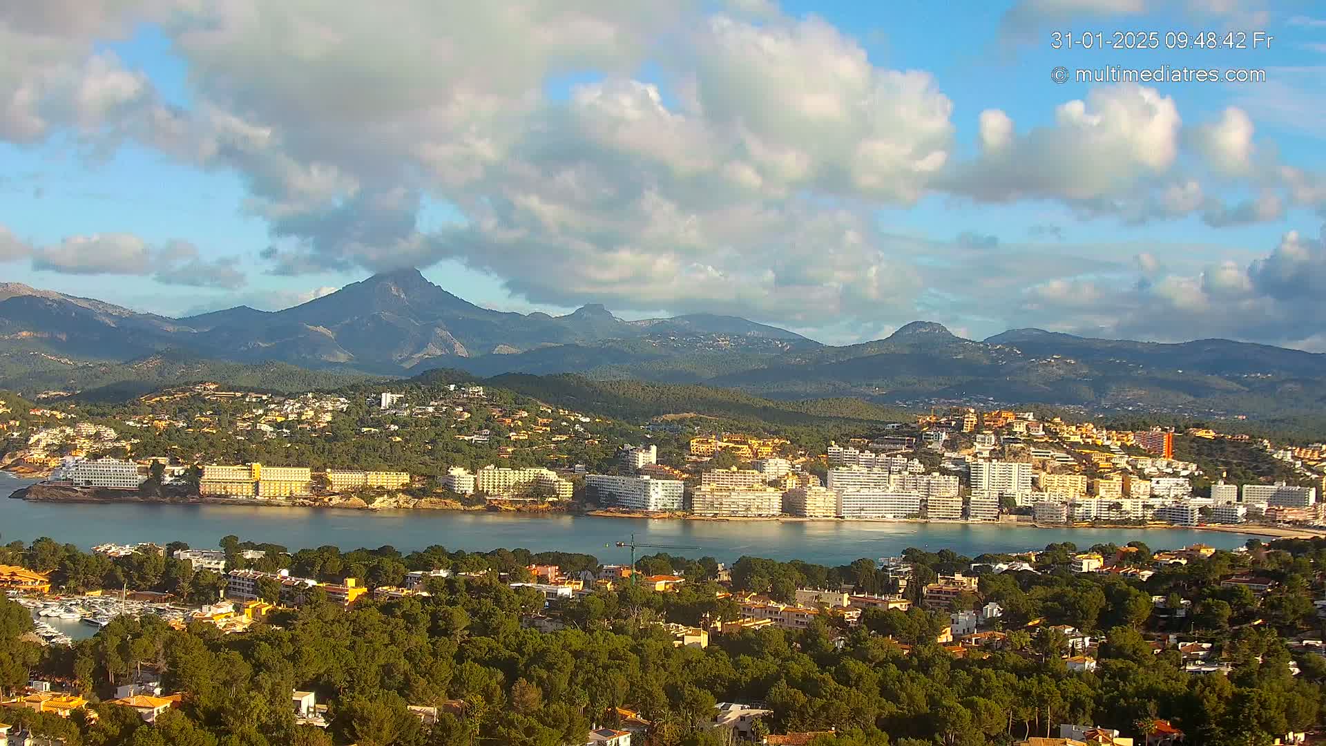A coastal town with numerous buildings nestles at the base of a mountain range under a partly cloudy sky.
