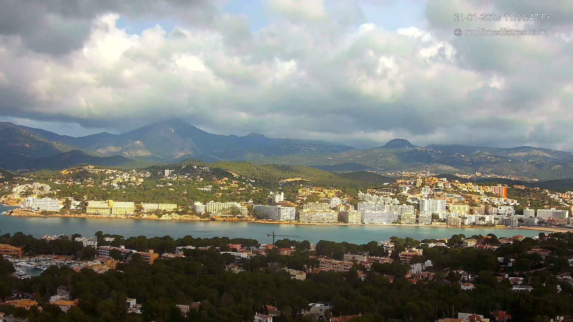 A coastal town nestled between a bay and mountains under a partly cloudy sky.