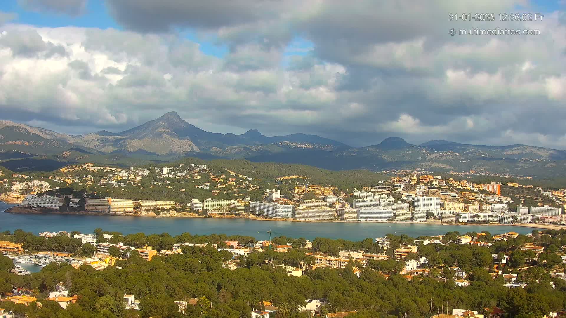 A coastal town nestled among green hills and mountains under a partly cloudy sky, with a calm body of water separating the town from a row of hotels.