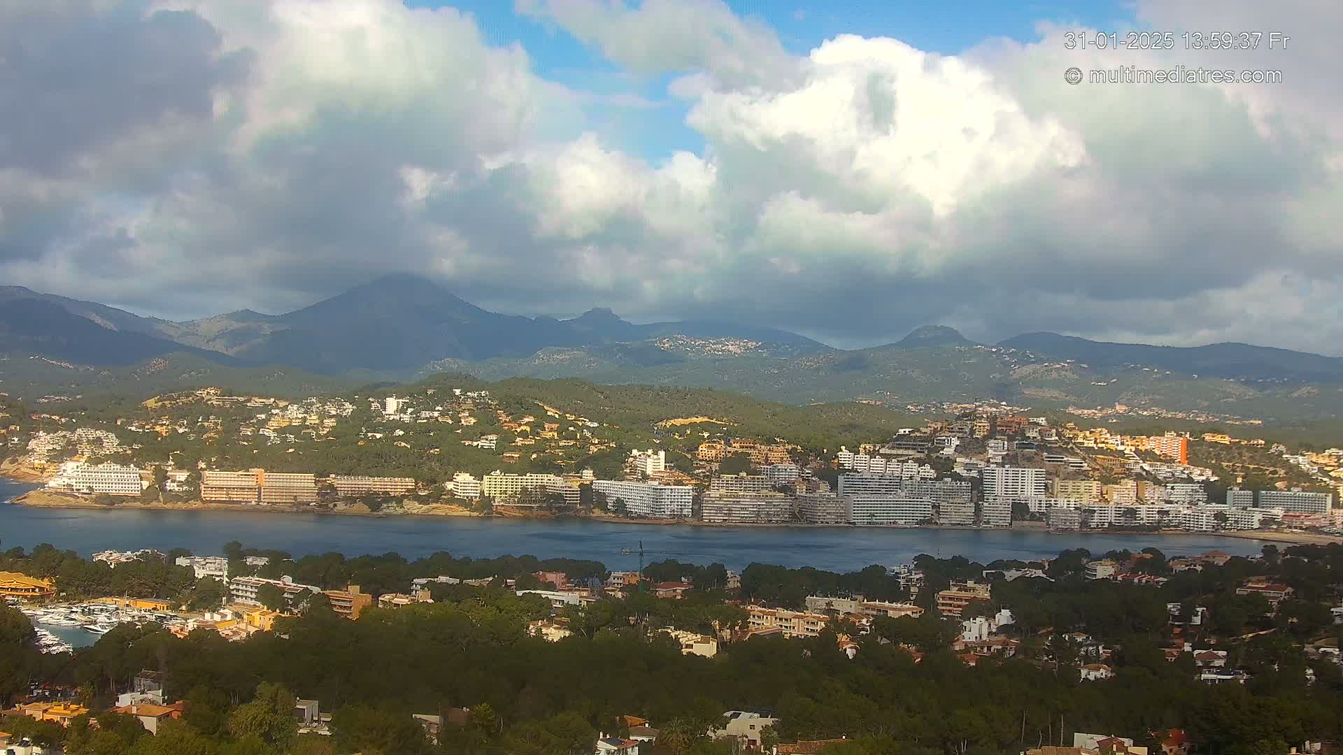 A coastal town with many buildings sits nestled between a body of water and a range of mountains under a partly cloudy sky.