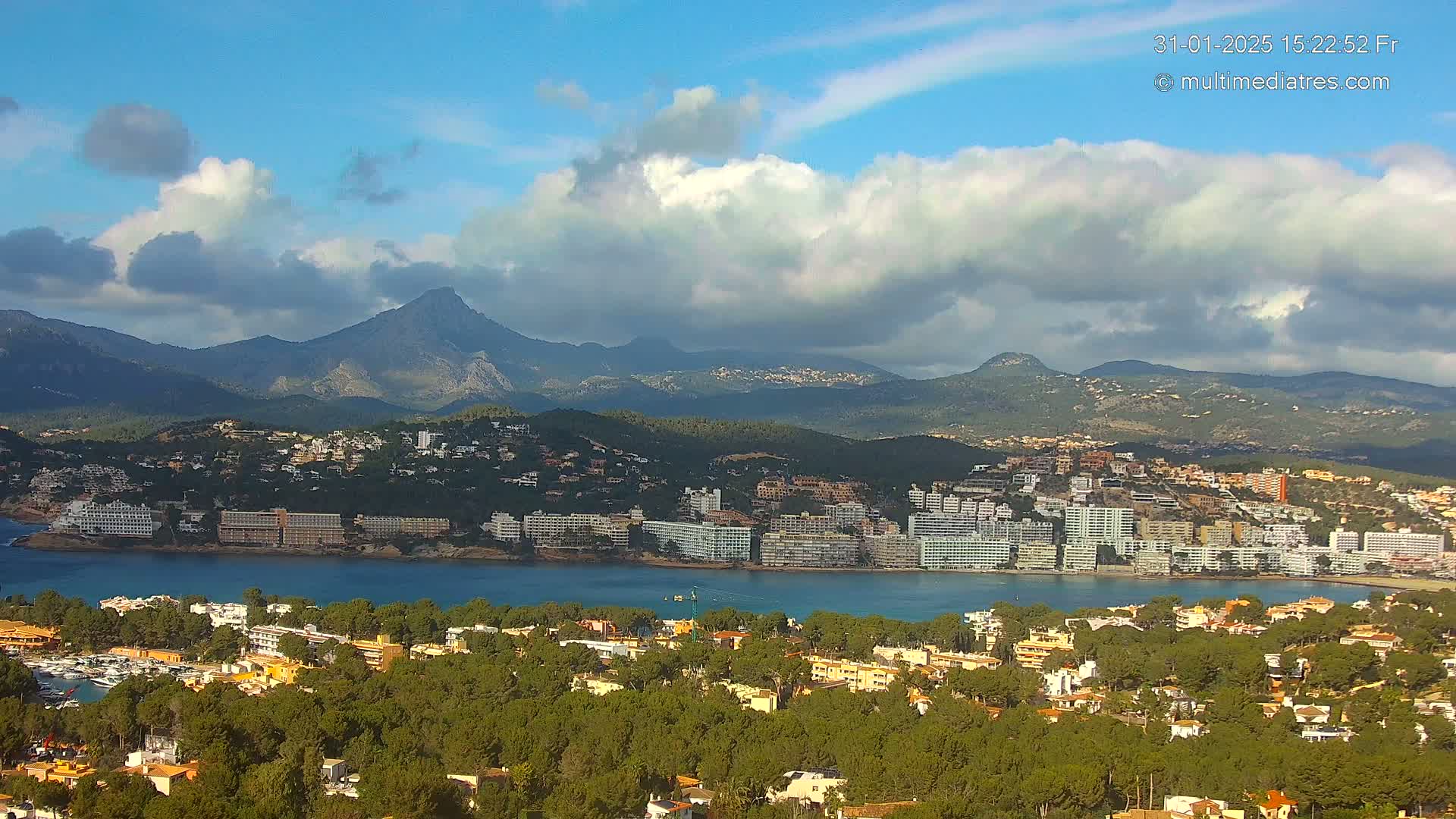 A coastal town nestled amongst green hills and mountains under a partly cloudy sky.