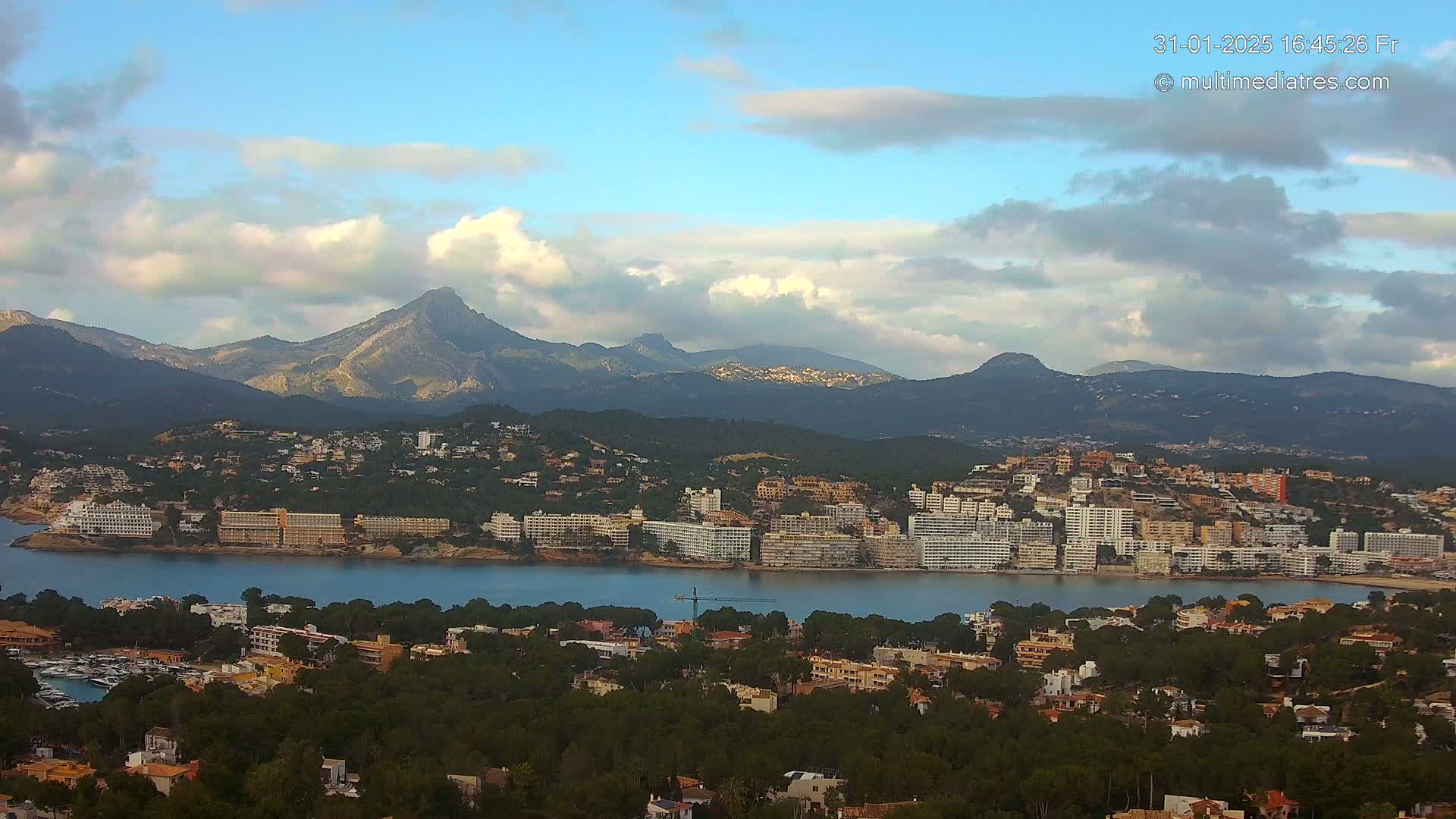 A coastal town nestled between a calm bay and a mountain range under a partly cloudy sky.