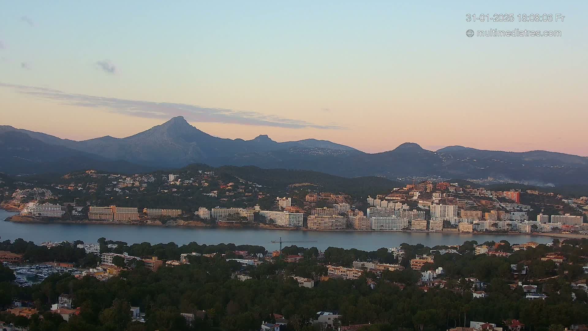A coastal town with numerous buildings nestled along a calm body of water, backed by a range of mountains under a soft, pastel sunset sky.
