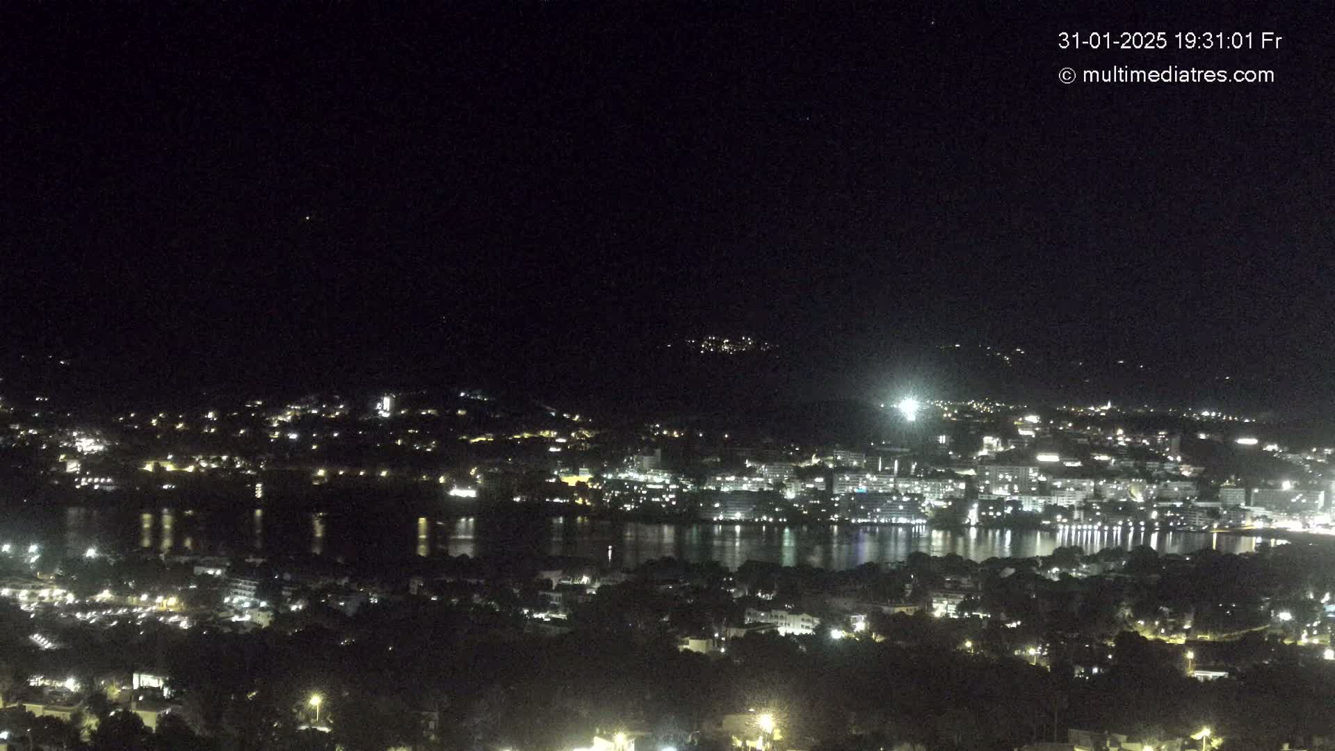 A nighttime, high-angle view shows a city illuminated by lights bordering a dark body of water, under a clear night sky.