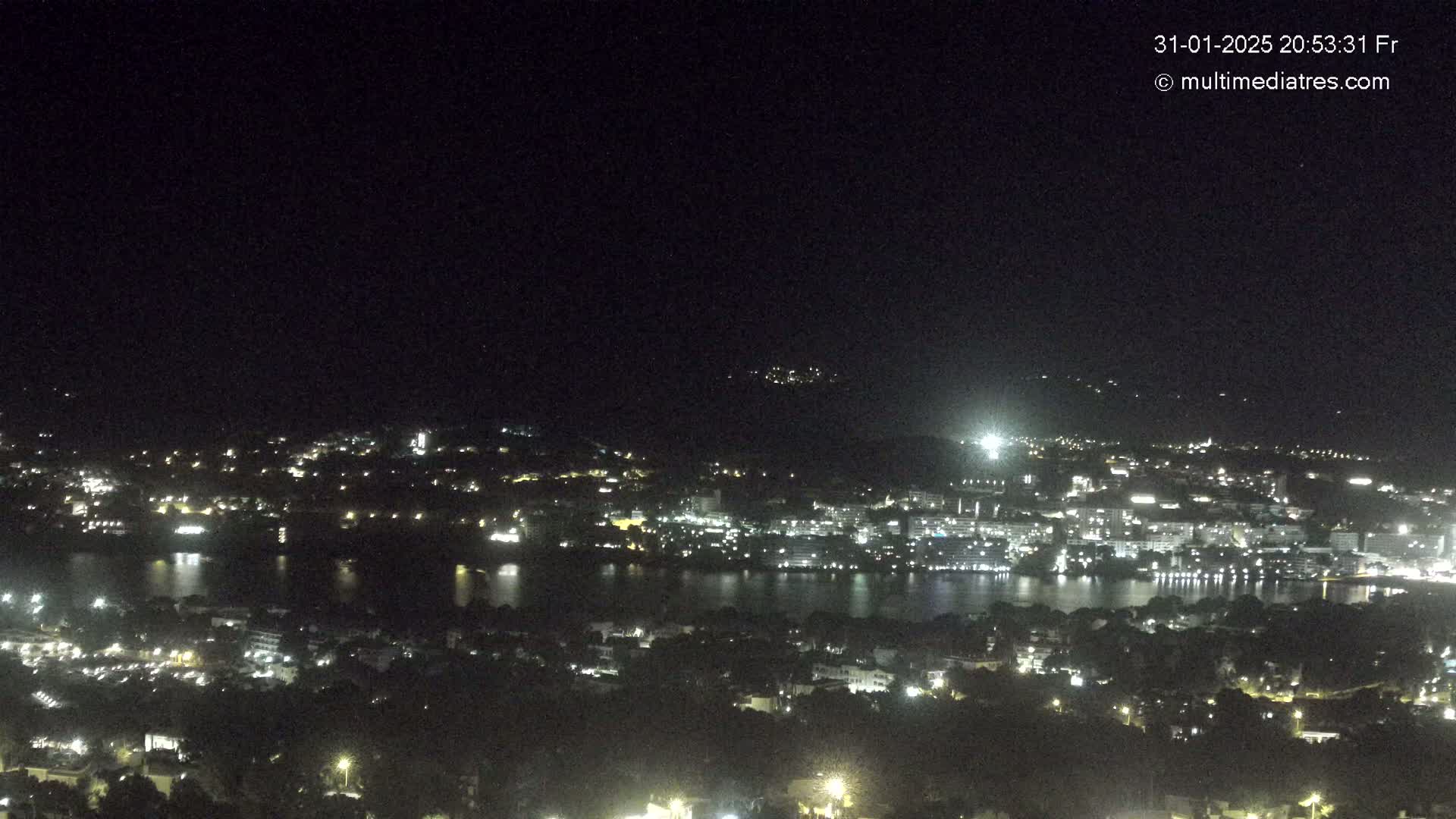 A nighttime, high-angle view shows a city with lights next to a dark body of water, all under a clear, dark sky.