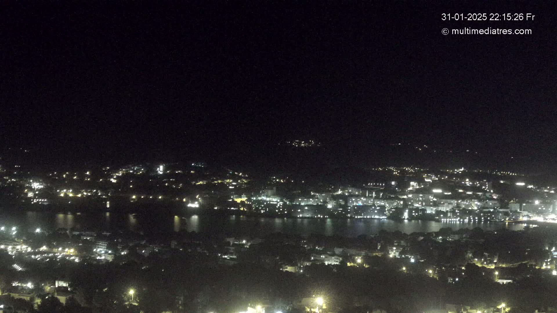 A nighttime, high-angle view shows a city with its lights illuminated beside a dark body of water, under a clear, dark sky.