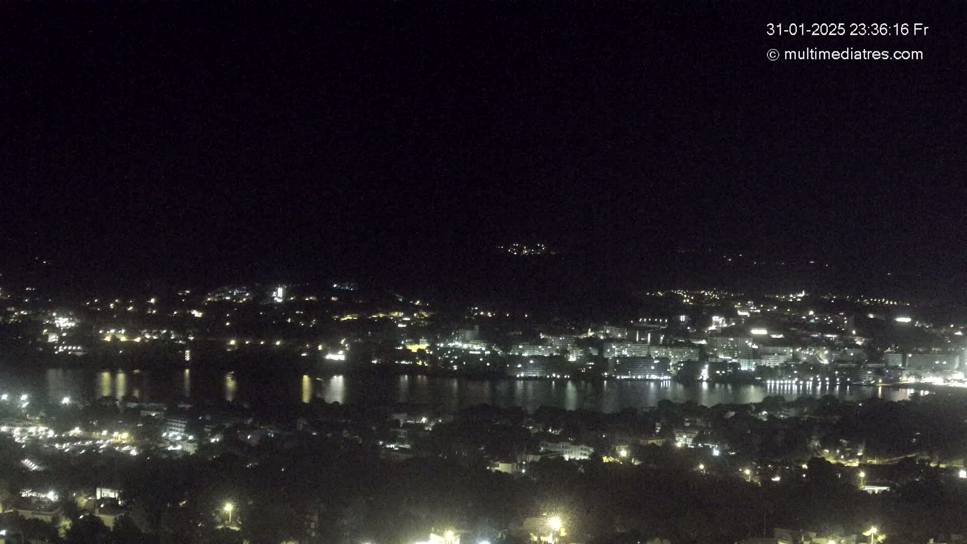 A nighttime view of a city illuminated by lights surrounding a body of dark water, under a clear, dark sky.