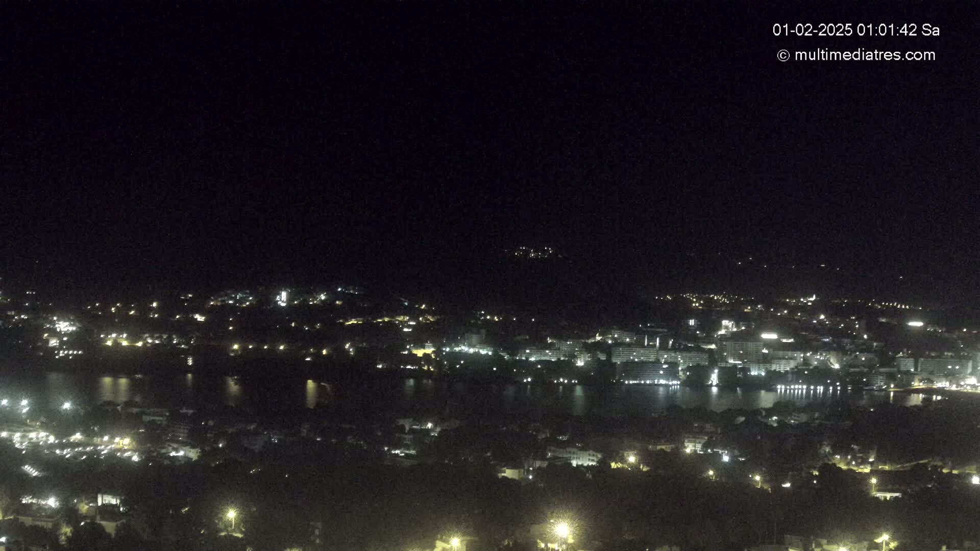 A nighttime, high-angle view shows a city spread across a dark body of water, illuminated by numerous lights.