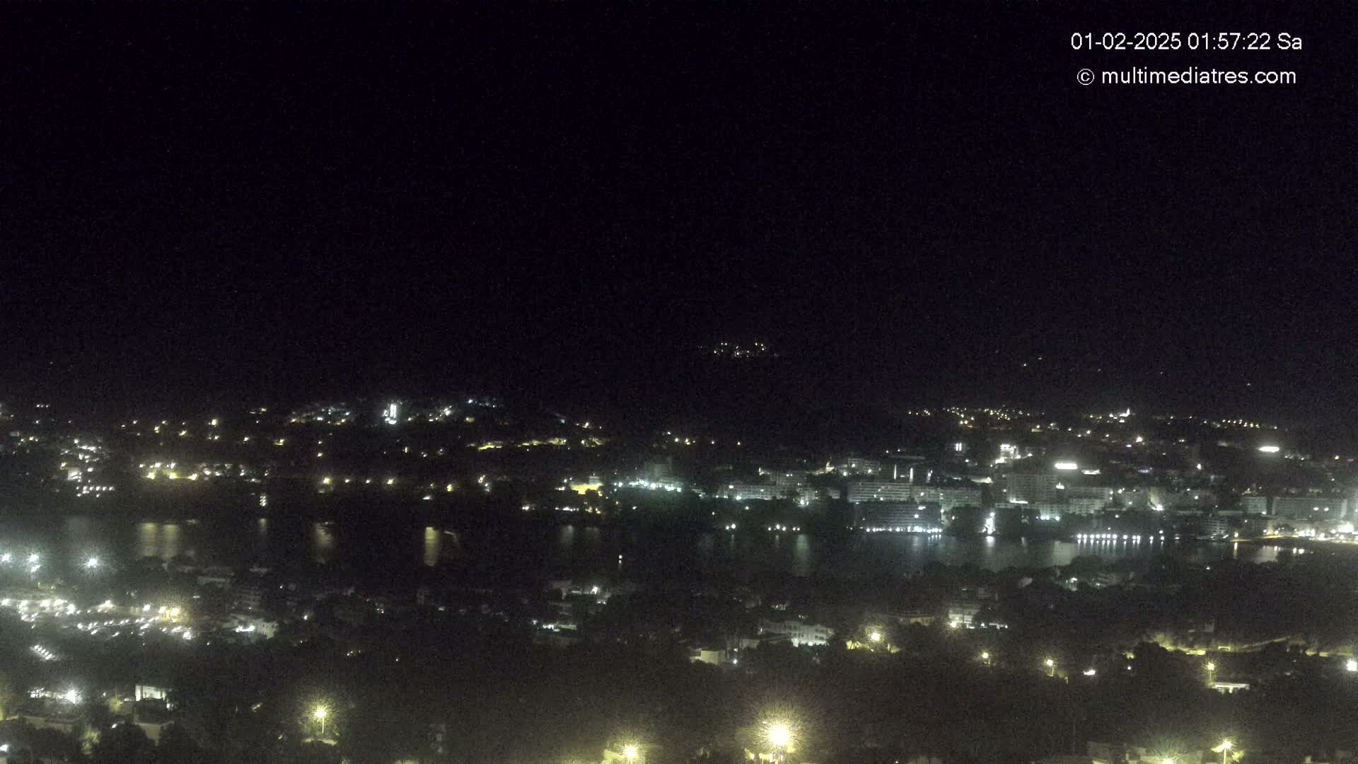 A nighttime, high-angle view shows a city and body of water with scattered lights illuminating buildings and streets under a clear, dark sky.