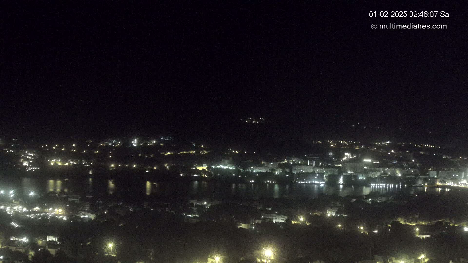 A nighttime, high-angle view of a city and body of water illuminated by lights, under a clear, dark sky.