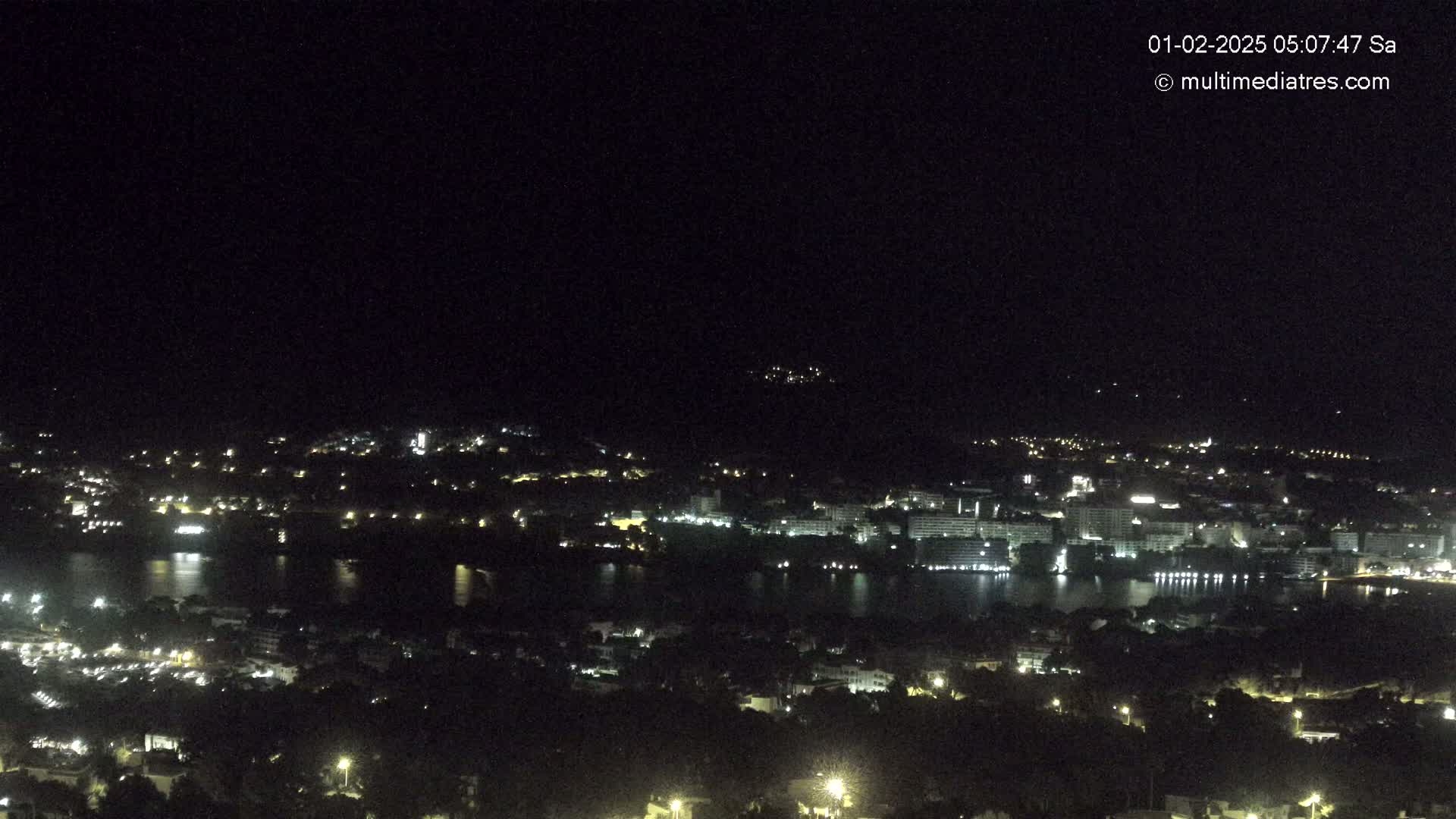 A nighttime, aerial view of a city with a body of water, showing scattered lights across buildings and hills.